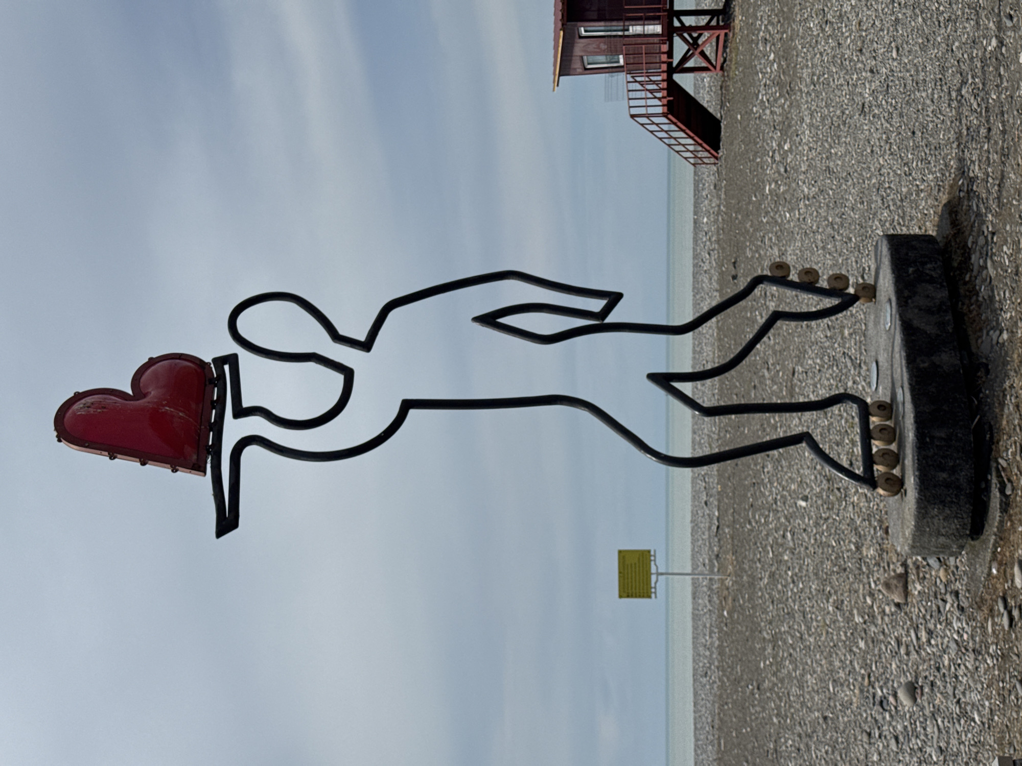 A medium shot depicts a black, wire sculpture of a man holding a red heart, set against a backdrop of a pebble beach, the Black Sea, and a pale blue sky in Batumi, Georgia. In the background, slightly above and to the right of the sculpture, stands a lifeguard tower with an exterior staircase. A small, yellow sign is located further in the background, near the sea. The sculpture is mounted on a dark-colored base and is positioned near the edge of the beach.