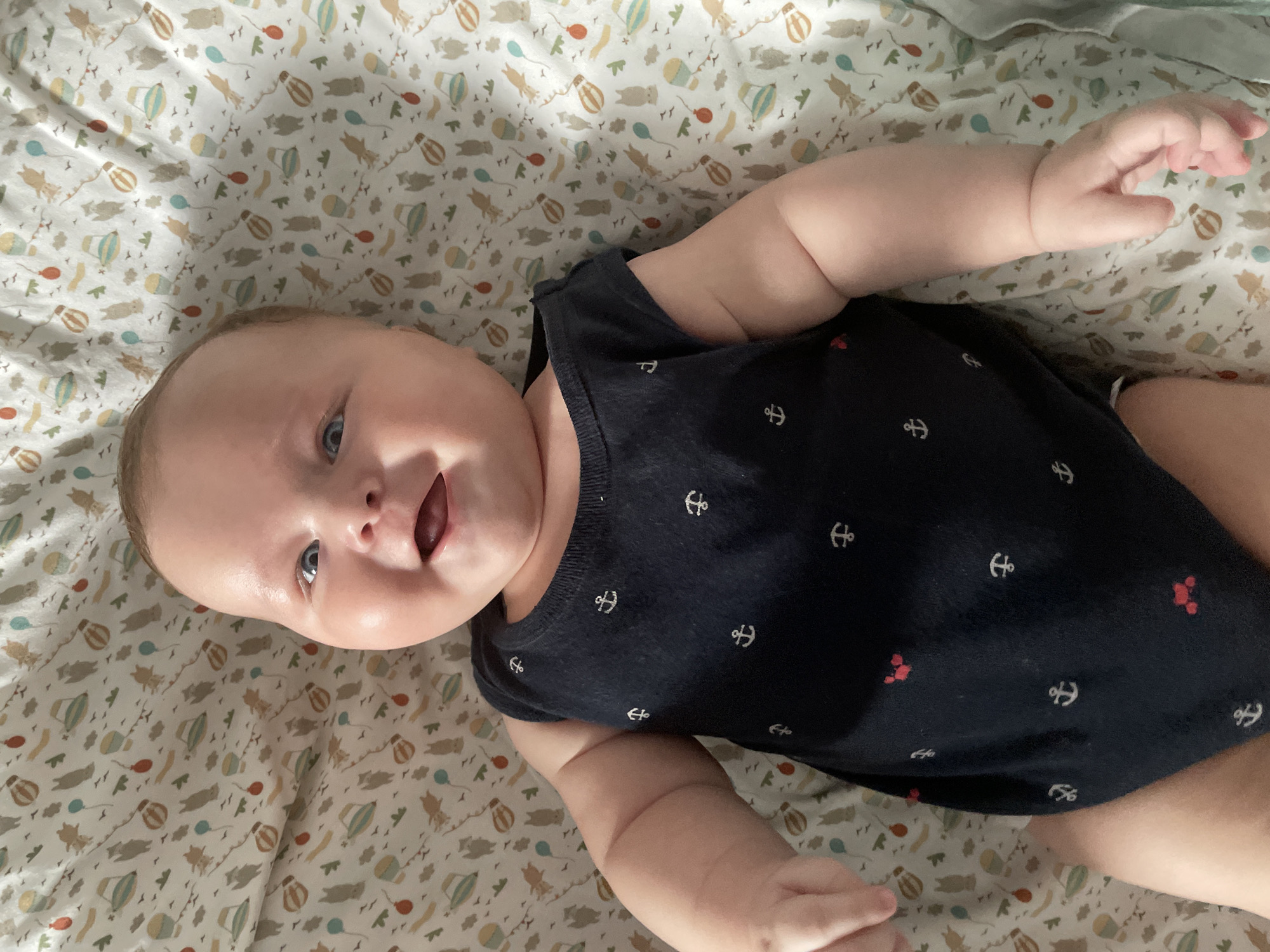 A high-angle, close-up shot shows a baby lying on its back on a patterned surface. The baby, facing the camera, has light skin, blue eyes, and rosy cheeks. Its mouth is open in a smile. The baby wears a dark blue sleeveless onesie adorned with white anchor patterns and a few small red crab designs. The background is a light-colored fabric with a repeating pattern of cartoonish hot air balloons and animals. There is no indication of Stavropol, Russia, in the image.