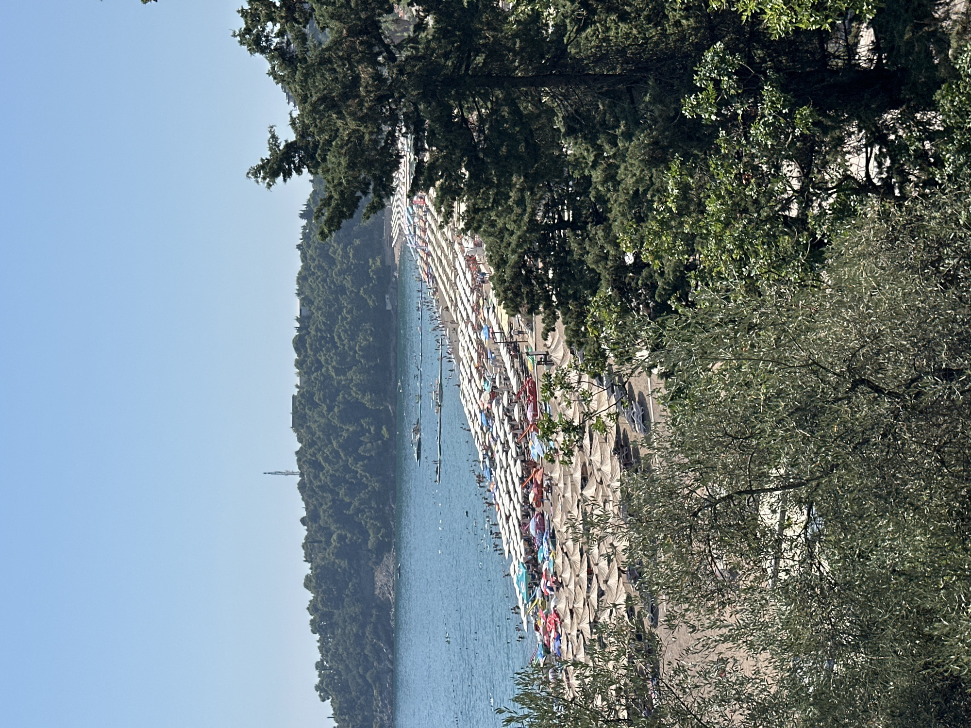 The image shows a crowded beach in Boreti, Montenegro. The beach is covered with numerous umbrellas and loungers, indicating a busy summer day. Many people are visible swimming in the clear blue sea or relaxing on the shore. The background features a lush green hillside dotted with trees, and a telecommunications tower can be seen on the summit. The water extends to the horizon, meeting a clear, bright blue sky. In the foreground, foliage from trees partially obscures the view of the beach. Several boats are anchored in the water, suggesting it's a popular spot for various water activities.