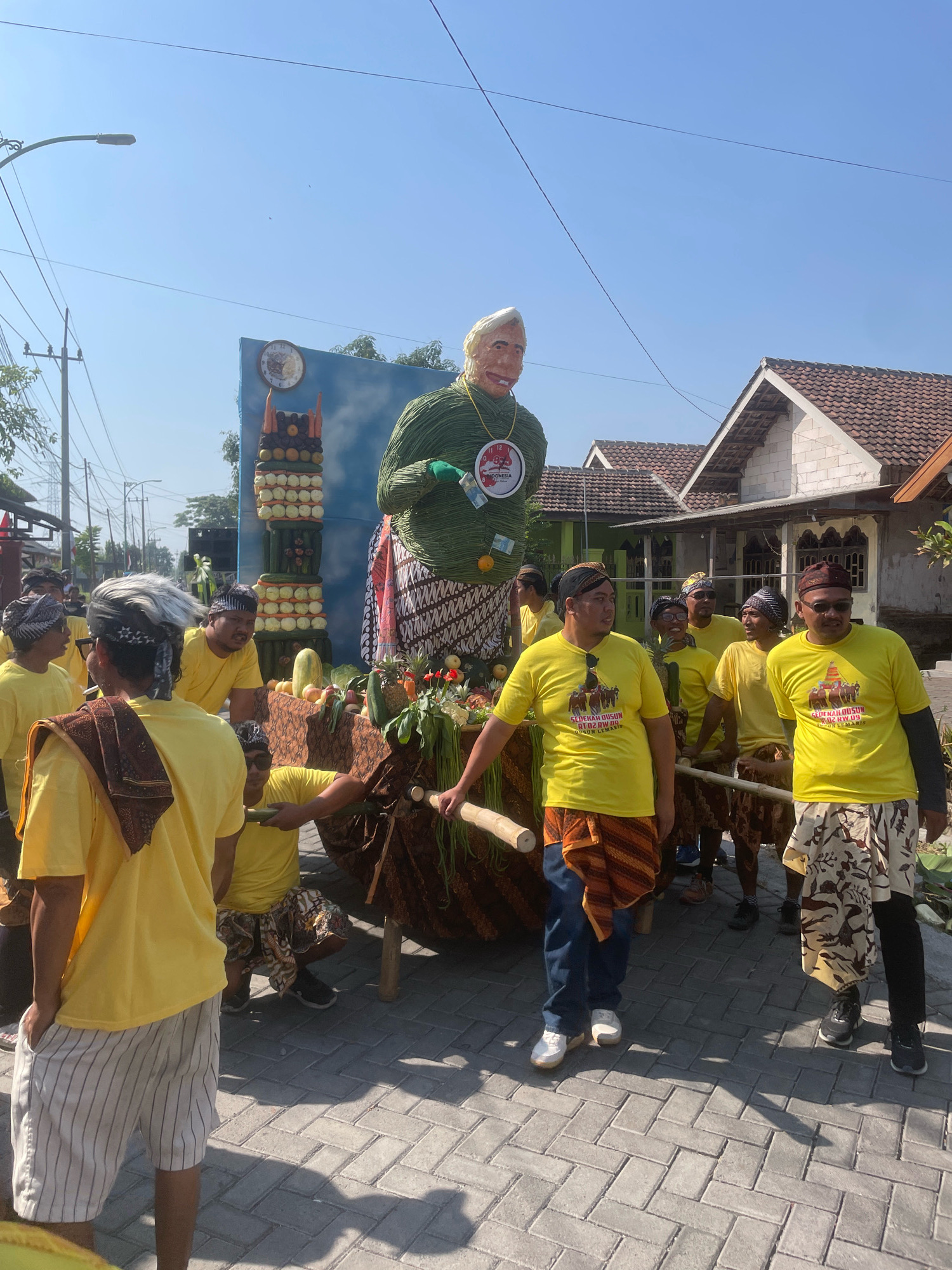 The image depicts a street procession in the city of Kebon Waris, Indonesia, likely during a festival or cultural event. A large, green, woven effigy resembling a person with blonde hair and a smiling face is prominently displayed on a cart. The effigy is adorned with a red badge. Beside it, a tall, stacked display of various fruits and vegetables, including oranges, apples, bananas, cucumbers, and what appear to be potatoes, is also part of the procession.
A group of men, all wearing bright yellow t-shirts and traditional Indonesian sarongs or patterned skirts, are actively involved in moving the cart. Some are carrying poles attached to the cart, indicating they are responsible for its transportation. Others stand by, observing or participating in the event. The men's attire suggests a collective identity or affiliation.
The environment shows a paved street with a cobblestone-like pattern. Houses with tiled roofs and white brick walls are visible in the background, characteristic of residential areas. Electrical wires and a lamppost suggest an urban or semi-urban setting. The sky is clear and blue, indicating fair weather, and shadows on the ground suggest a sunny day. The overall scene conveys a sense of community engagement and cultural celebration.
