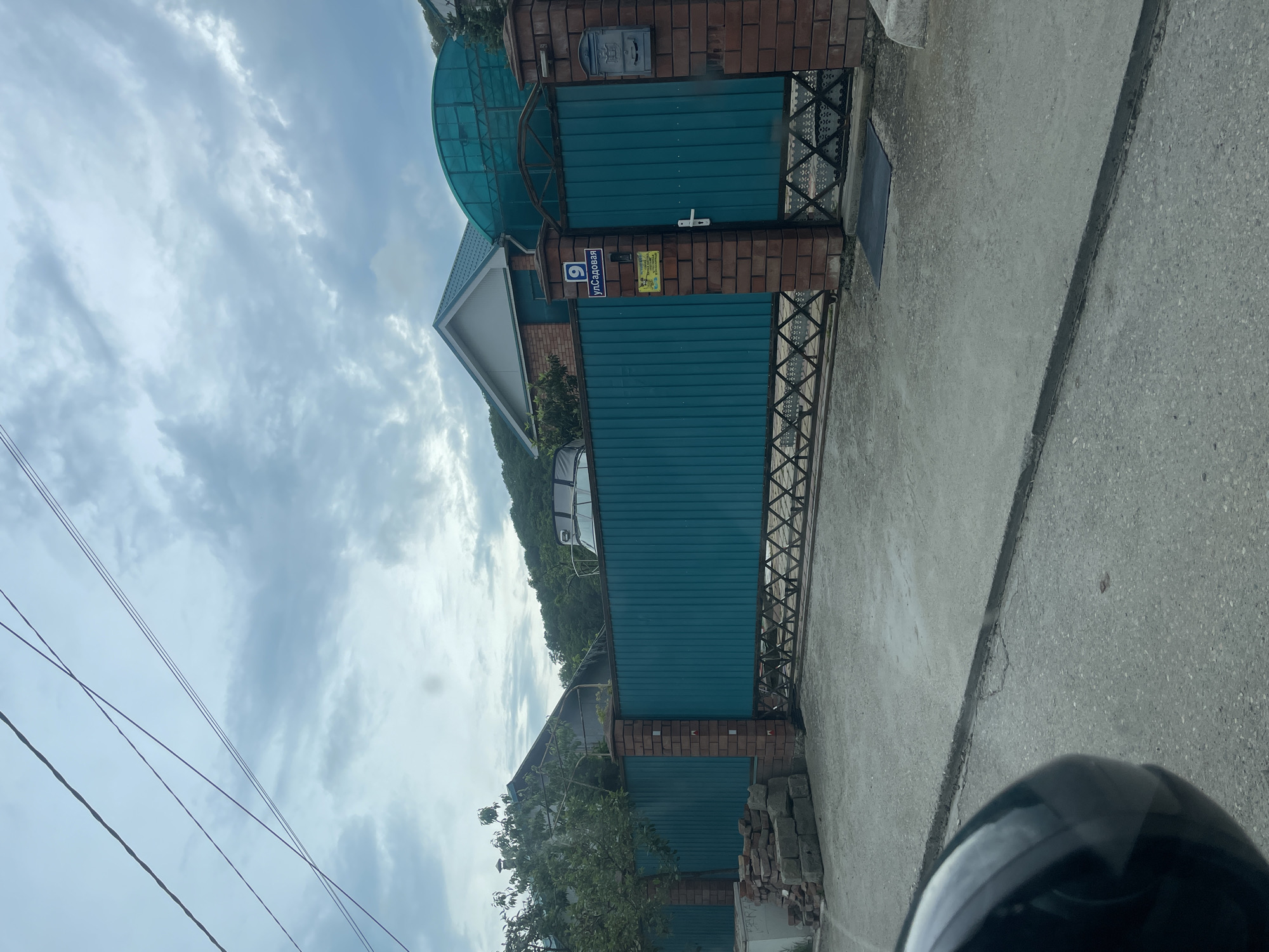 A medium, slightly angled, outdoor shot shows a property entrance in Olginka, Russia. A blue address plate with "ул.Садовая 9" is affixed to the brick column of the gate. The gate is made of blue corrugated metal. A transparent, arched roof extends over the gate's pedestrian entrance. A mailbox is visible above the pedestrian entrance. Bricks are stacked on the ground next to the gate. Power lines run in the top left corner of the image, and a portion of a helmet is visible in the bottom right. The sky is overcast.