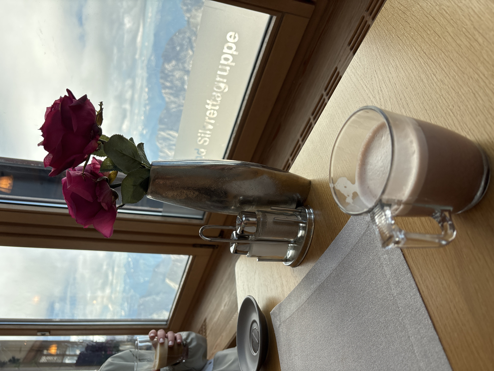 The image shows a table setting in what appears to be a restaurant or cafe with a panoramic view of mountains through a large window. In the foreground, a glass mug containing a frothy, chocolate-colored beverage sits on a wooden table. To the left, a grey placemat covers part of the table, and a small, dark grey saucer is visible. A salt and pepper shaker set with a metal rack is positioned to the right of the placemat.
In the middle ground, a metallic vase holds two artificial pink roses. The background is dominated by a large window offering a breathtaking view of snow-capped mountains under a cloudy sky. A white decal with text, partially obscured, reads "Silvrettagruppe," suggesting the location is likely in the Silvretta region, potentially near Ehrwald, Austria, given the mountainous terrain.
A person's hand is visible on the far left, holding a glass of a similar beverage. The overall atmosphere suggests a cozy and scenic dining experience.
