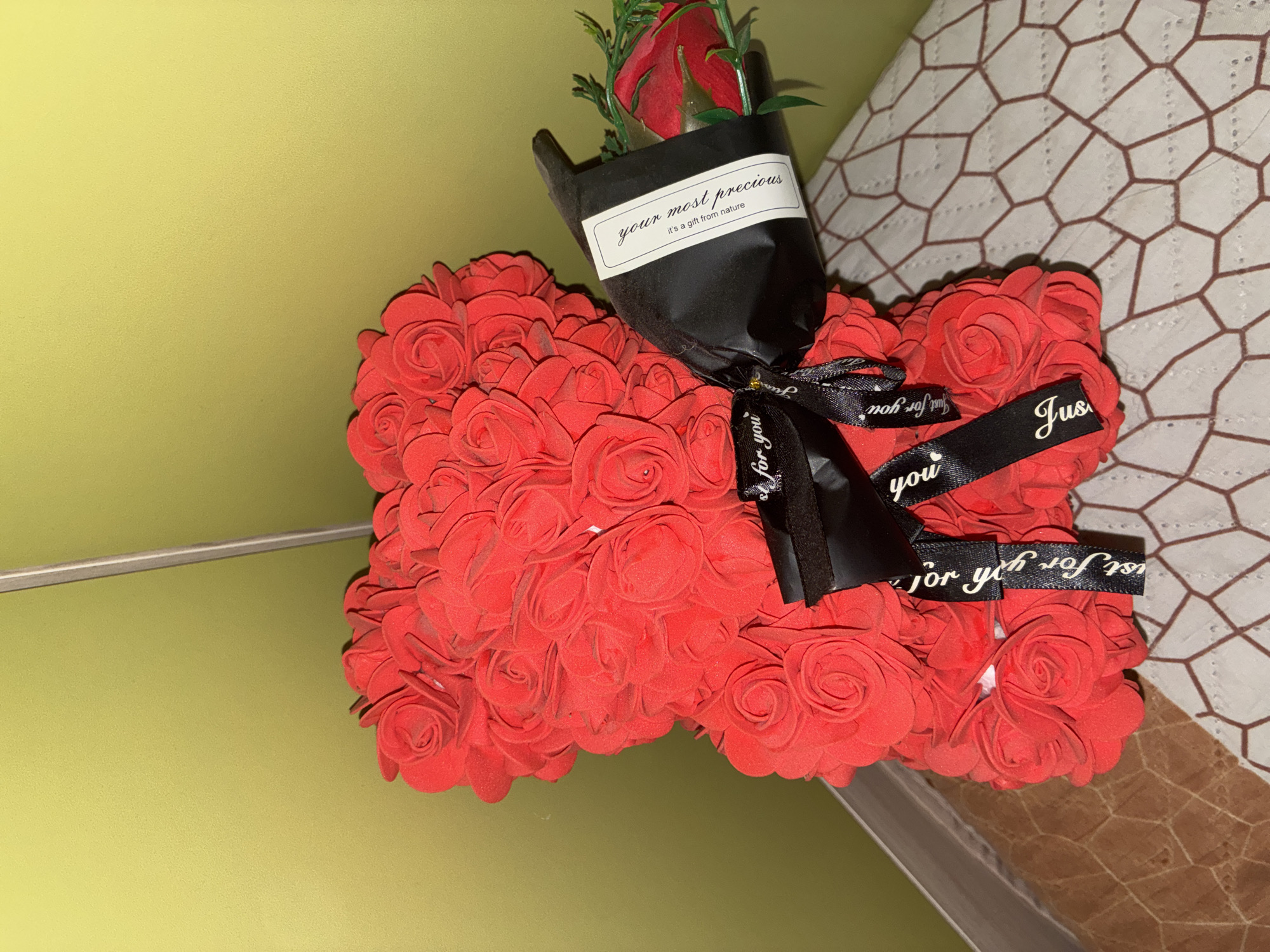 A red rose bear is on display, set against a plain, muted lime green wall. The bear, a voluminous figure constructed from numerous small, individual red roses, stands prominently. A black ribbon, adorned with white lettering that reads "Just for you," is tied around the bear's midsection. Attached to the bear is a bouquet of one red rose, wrapped in black paper, with a small white label that reads "your most precious, it's a gift from nature." The bear and bouquet rest on a surface covered by a patterned textile, which has a geometric design of interconnected hexagons in white and brown. The lighting in the scene is moderately bright, casting some soft shadows. The image captures a still life arrangement, with no people present and no discernible urban or specific geographical context.