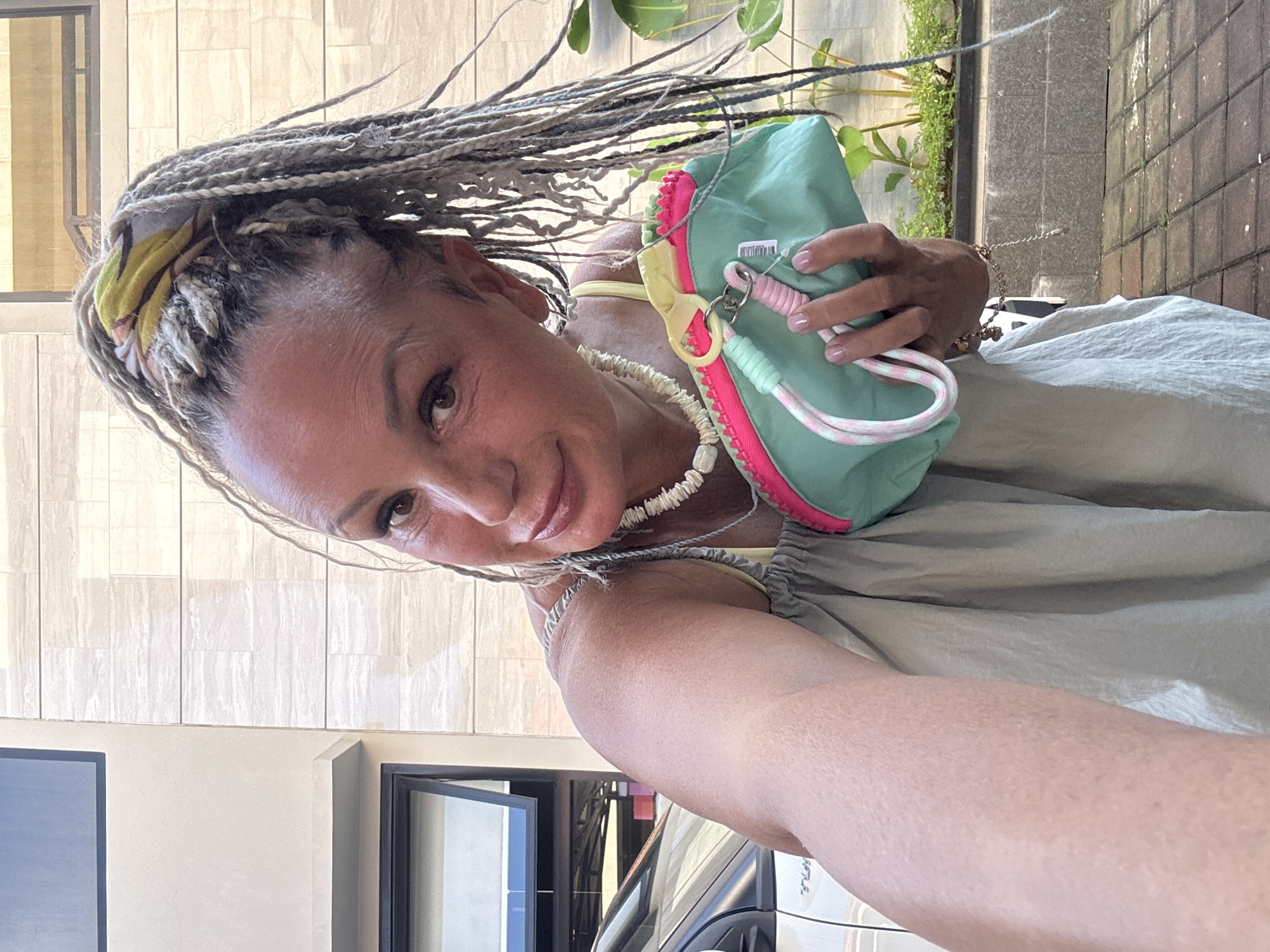 The image shows a woman taking a selfie. She has braided hair with a colorful scarf and is wearing a necklace. She is holding a light green pouch with a pink zipper. In the background, there is a building with light-colored tiles and a window. There is also greenery and a glimpse of a car. The exact location within Kuta, Indonesia, cannot be determined from the visual information provided.