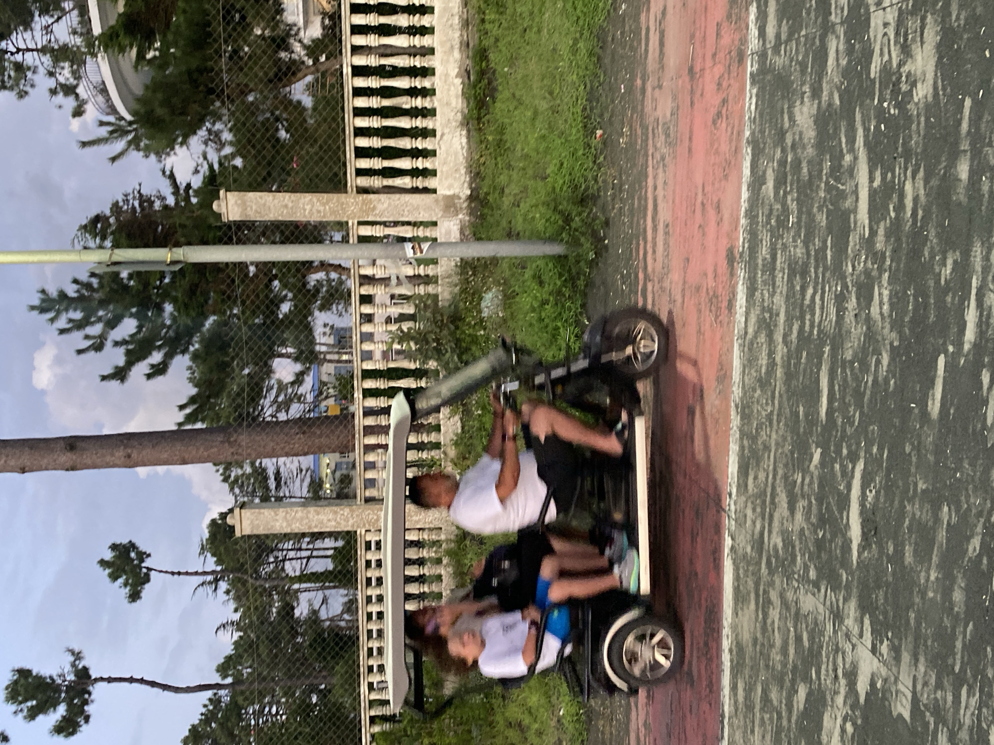 A family is riding in a black golf cart. The father is driving, wearing a white t-shirt and black shorts. His wife is in the passenger seat, talking on her phone. Their son is in the back, wearing a white t-shirt and blue shorts. The golf cart is on a red and grey paved path. In the background, there is a white fence with ornate columns, lush green grass, and tall pine trees. Behind the trees, a large, white building with a balcony can be seen. The sky is cloudy. The scene is likely in Ureki, Georgia, known for its beaches and pine forests.