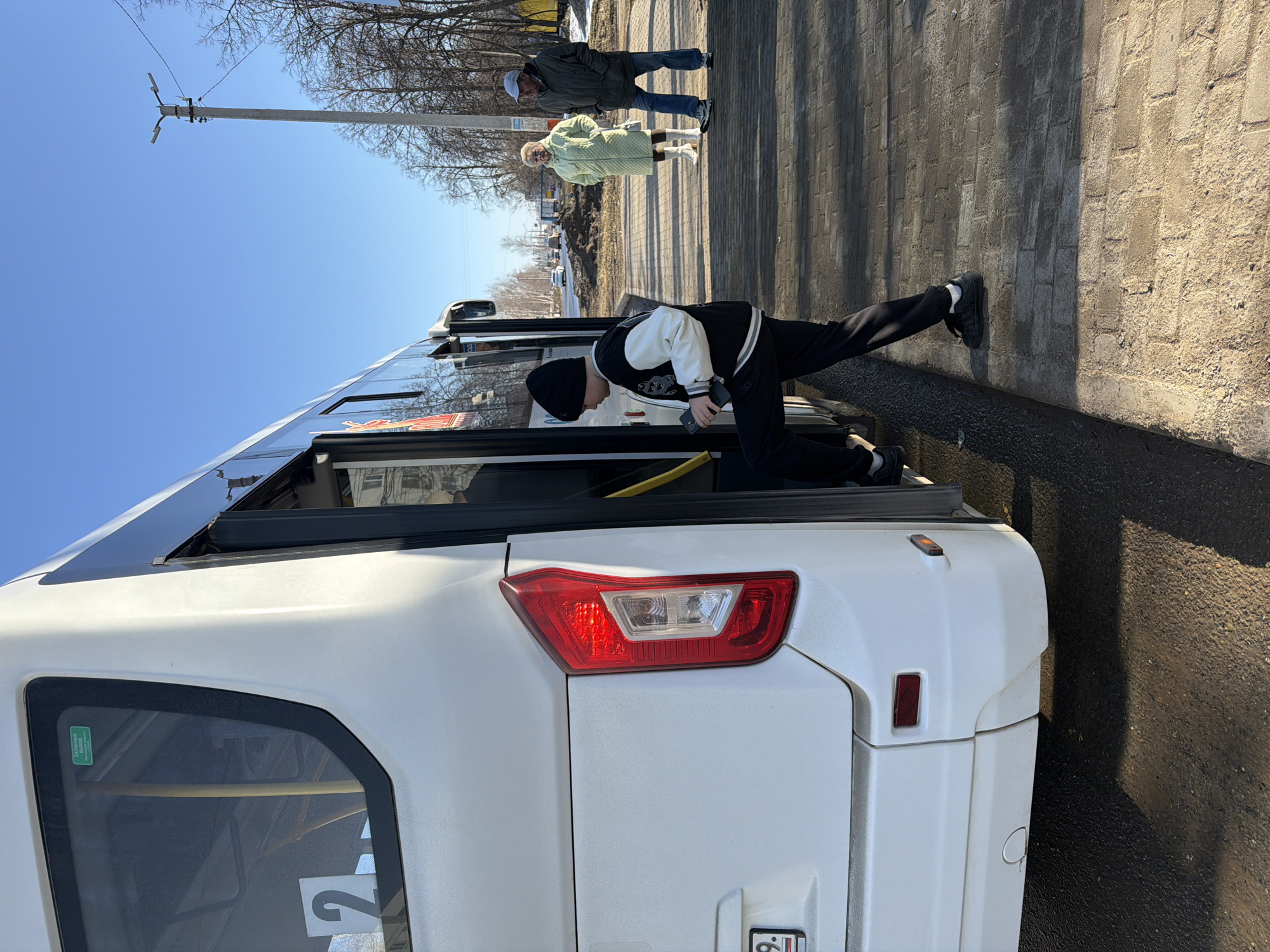 A young person wearing a black beanie, black pants, and a black and white jacket is leaning into the open rear door of a white bus parked on a paved road. The person's body is angled as they reach inside the bus with a yellow handle visible in the doorway. Two other people are walking on a sidewalk in the background. A utility pole is on the left. The photo was taken in Chaikovsky, Russia.