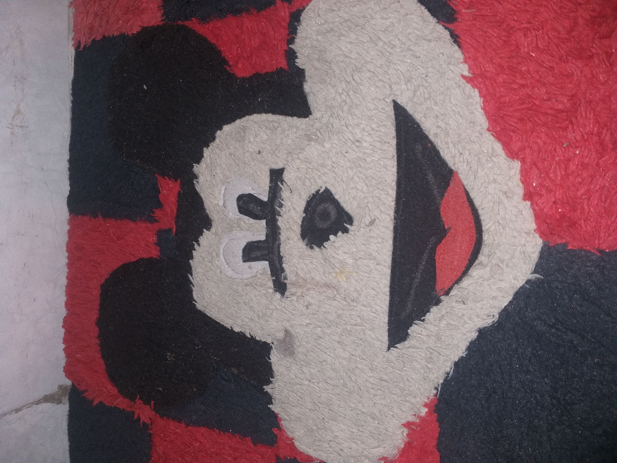 The image shows a close-up of a rug with a Mickey Mouse design. The rug has a checkered pattern of red and black, with a large white face of Mickey Mouse in the center. The face has black eyes, a black nose, and a red mouth with white teeth. The rug appears to be placed on a tiled floor. The lighting is dim, and there are some dust particles visible on the rug. The background is a wall with a textured pattern. The image is undated and unlocated.