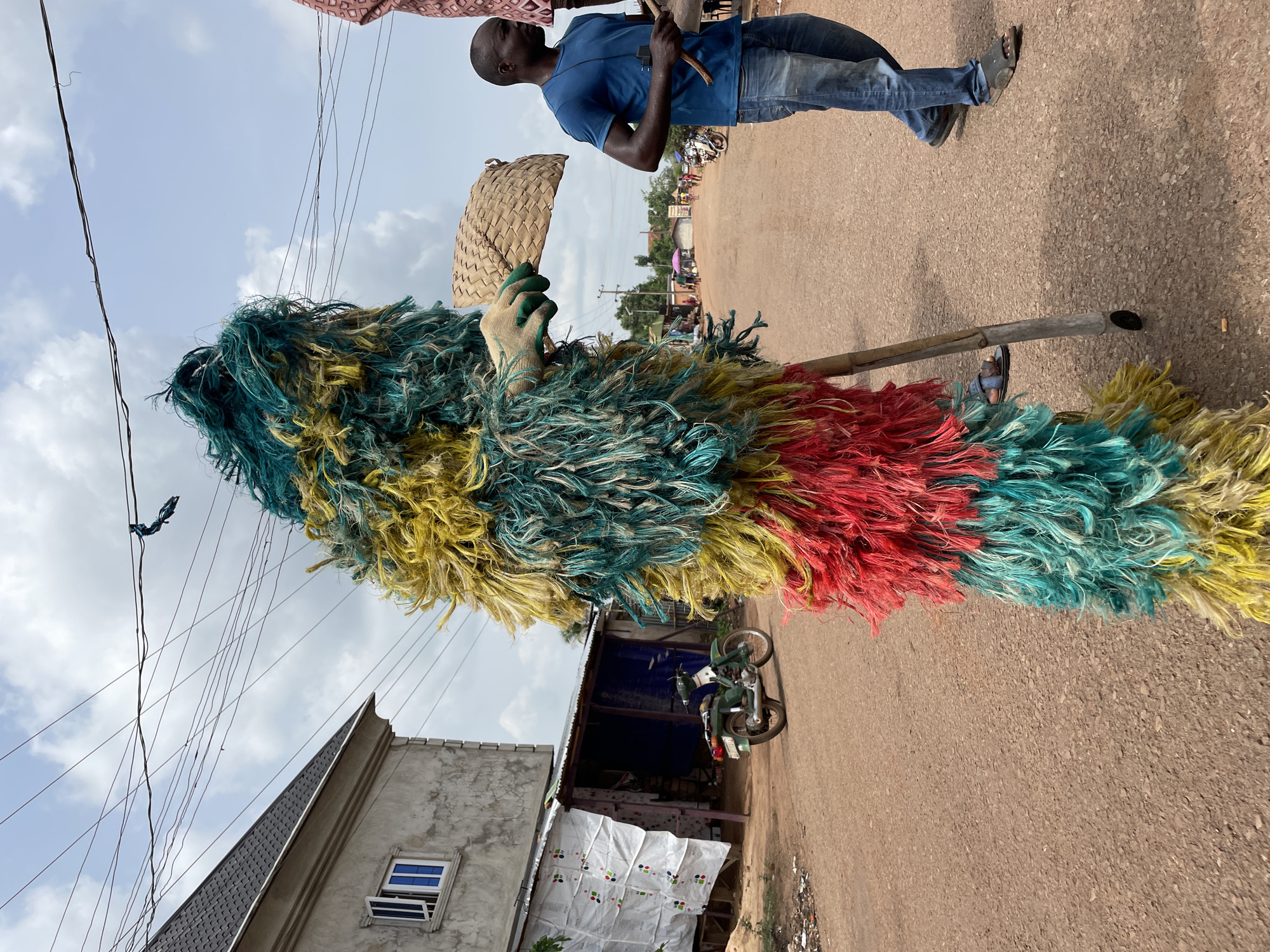 In Agba Umana, Nigeria, a colorful raffia masquerade costume is centered on a brown dirt road. The costume displays horizontal bands of yellow, green, and red raffia. A gloved hand holds a woven fan. A wooden pole acts as a brace, with a person's foot in a sandal visible at the base. To the left, a partially constructed building with a gray exterior, white-framed window, and a dark gray roof is visible. A green motorcycle is parked in front of a building shrouded in white and blue tarp. In the background, a man in a blue shirt and jeans is standing, holding a wooden object. The sky is overcast with visible power lines.