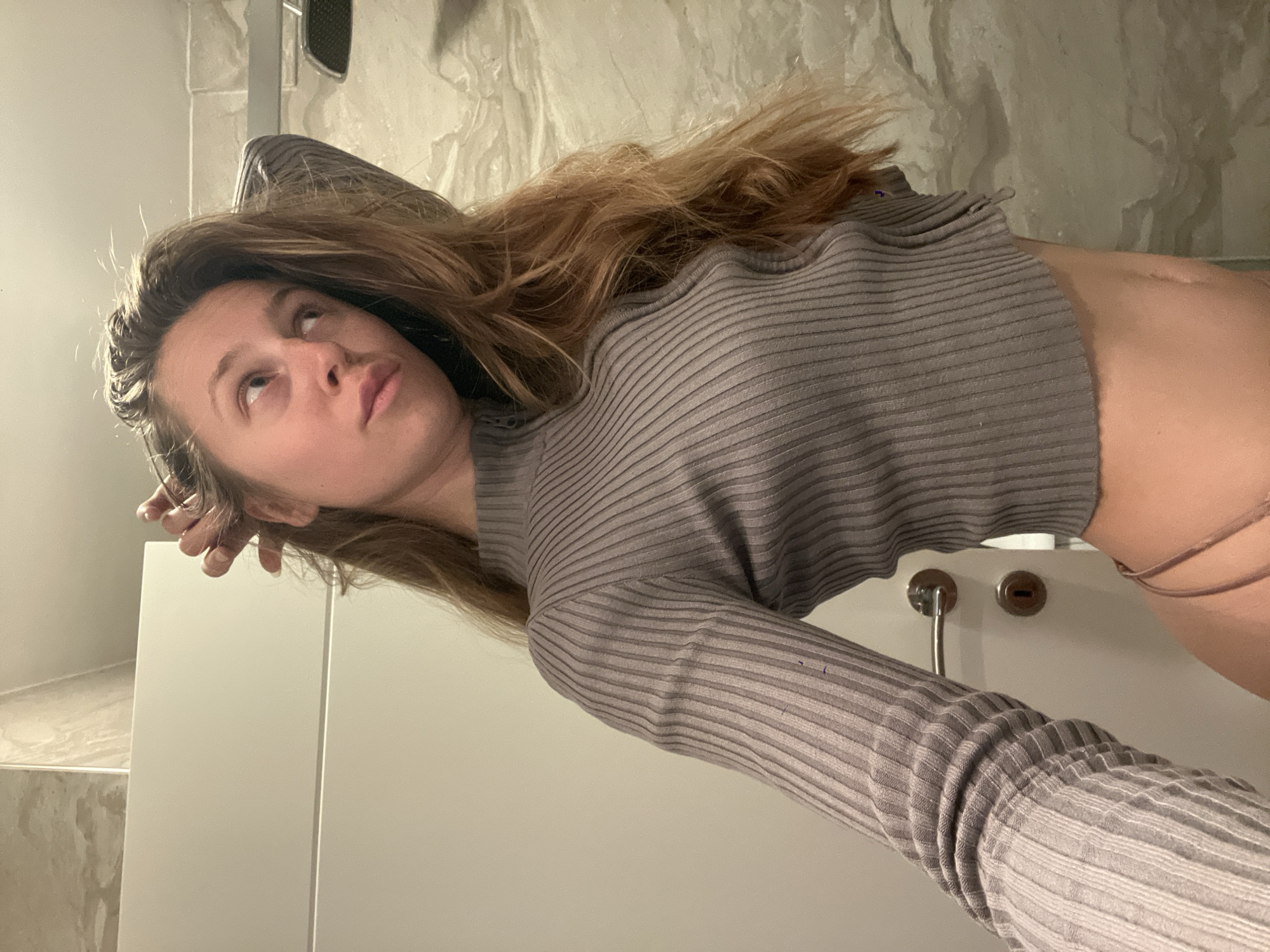 A young woman stands indoors, in what appears to be a bathroom. She wears a long-sleeved, ribbed gray shirt and a thin strap of what appears to be underwear is visible near her right hip. Her long brown hair is partially spread behind her. She is looking upwards with a somewhat contemplative expression. Her right hand is raised near her head, possibly touching her hair, while her left arm is stretched out in front of her, slightly bent at the elbow. The bathroom features a marble wall behind her and a white cabinet with a chrome handle and a keyhole in front of her. There is no immediately discernible evidence that would place the image in Bečići, Montenegro.