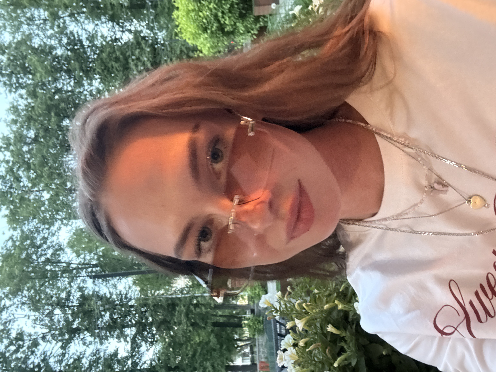 A close-up, eye-level selfie-style photograph depicts a young adult female outdoors in the city of Perm, Russia. The woman has long, wavy reddish-brown hair and is wearing rimless, light brown-tinted sunglasses. Her face is illuminated by a warm, golden light source from the right, casting a distinct shadow across her nose and left cheek. She exhibits a subtle, closed-mouth smile. She wears a white short-sleeved t-shirt with a dark red cursive design partially visible on the left chest, reading "Swe...". Multiple layered necklaces adorn her neck, including a thin silver chain, a longer silver chain with a heart-shaped pendant, and another lighter-colored beaded or chain necklace. In the lower left foreground, a bush of white trumpet-shaped flowers and green foliage is prominently featured, with portions of a dark outdoor table and chairs visible behind them. The background consists of dense green foliage from numerous deciduous trees under a light blue sky, indicating an outdoor park or garden setting. A faint utility pole is visible amidst the trees in the upper middle-left.