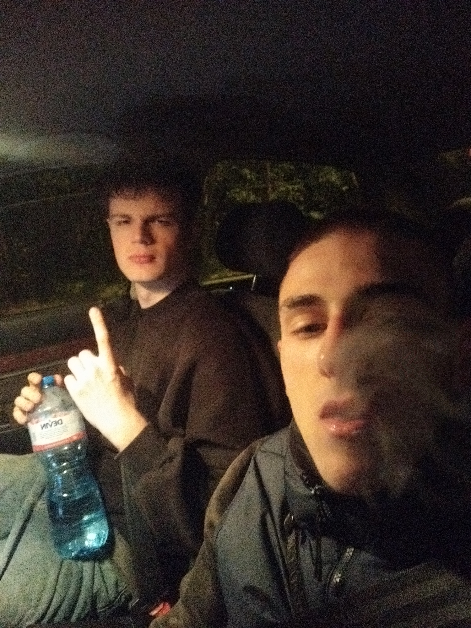 Two young males are in the front seats of a car. The male in the foreground is exhaling smoke, and wearing a dark jacket. The male in the background is holding a bottle of water, with the word "DEVIN" visible on the label, and holding up one finger. The car's interior is dark, and what appears to be trees are visible through the car window. The location cannot be confirmed in Sofia, Bulgaria.