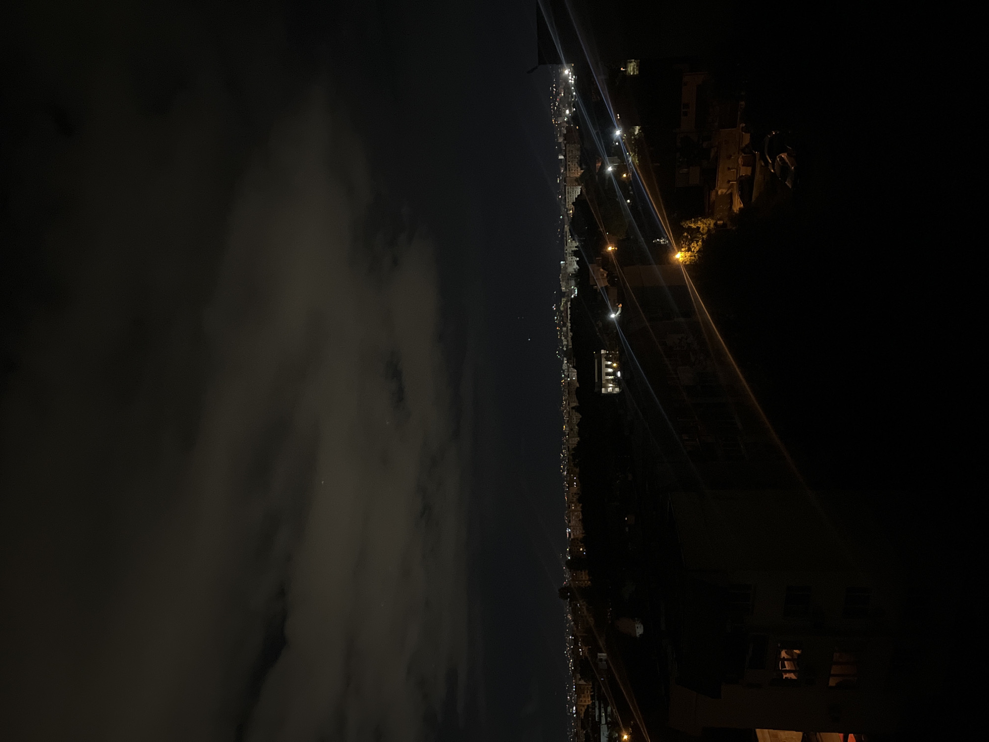 The image depicts a nighttime cityscape, possibly Sofia, Bulgaria, given the user's prompt. The left side of the image shows a cloudy sky, while the right side reveals an urban scene illuminated by streetlights and building lights. There are no discernible people or specific actions visible. The scene is dark, with the city lights providing the primary source of illumination. The environment appears to be an urban area with buildings and infrastructure. The picture is taken from a high angle.