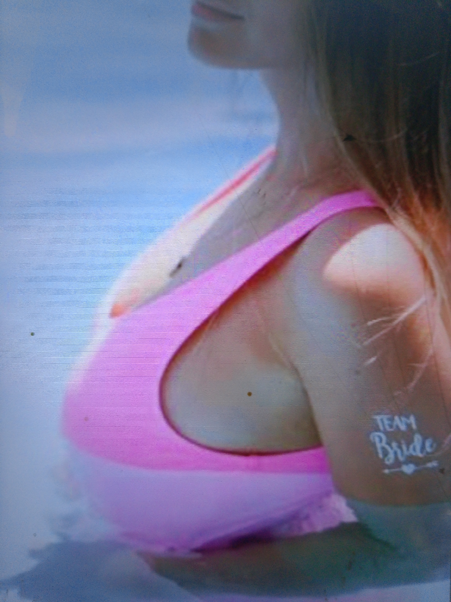 A woman with blonde hair and fair skin is partially submerged in blue water. She is wearing a pink swimsuit with a dark band around the top. A temporary tattoo on her upper arm reads "TEAM Bride" with two small hearts below. The image is blurry and grainy.