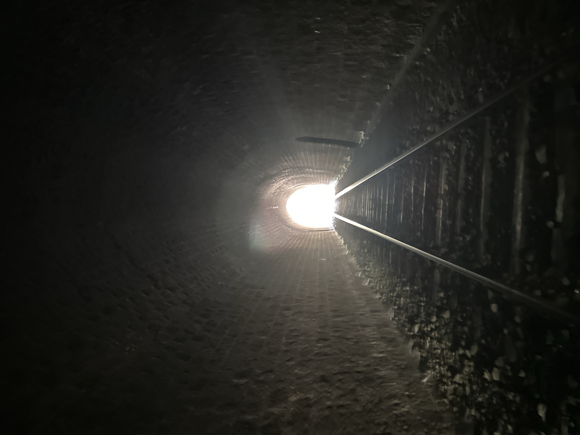 The image captures the interior of a dark brick tunnel with railway tracks running through it. The tunnel walls are constructed from aged, rough-hewn bricks, and the ground beside the tracks is covered in gravel and debris. At the end of the tunnel, a bright, white light emanates, suggesting an exit. The perspective is from a low angle, looking down the length of the tracks towards the light. There are no people or animals visible in the frame. The tunnel is located in Générargues, France, specifically the Pont-du-Gard railway tunnel, which is a disused railway tunnel.