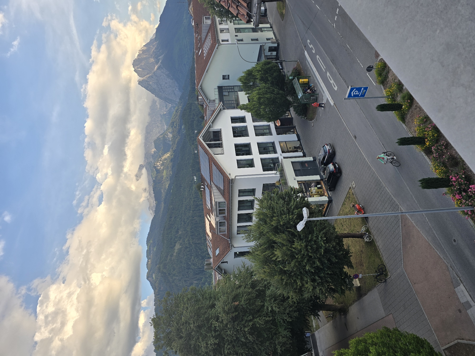 This image depicts a street scene in Garmisch-Partenkirchen, Germany, with a backdrop of mountains and clouds. In the foreground, a road curves to the right, with a cyclist riding on the bike path. To the left of the road are buildings with retail spaces on the ground floor, one of which displays "Performance" in its window. Further back, a bus stop is visible with people waiting. Behind the buildings, trees are abundant, leading up to a forested mountainside. The sky is partly cloudy, with sunlight illuminating the clouds and the mountain peaks in the distance. A parked red scooter and bicycles are also visible on the sidewalk.