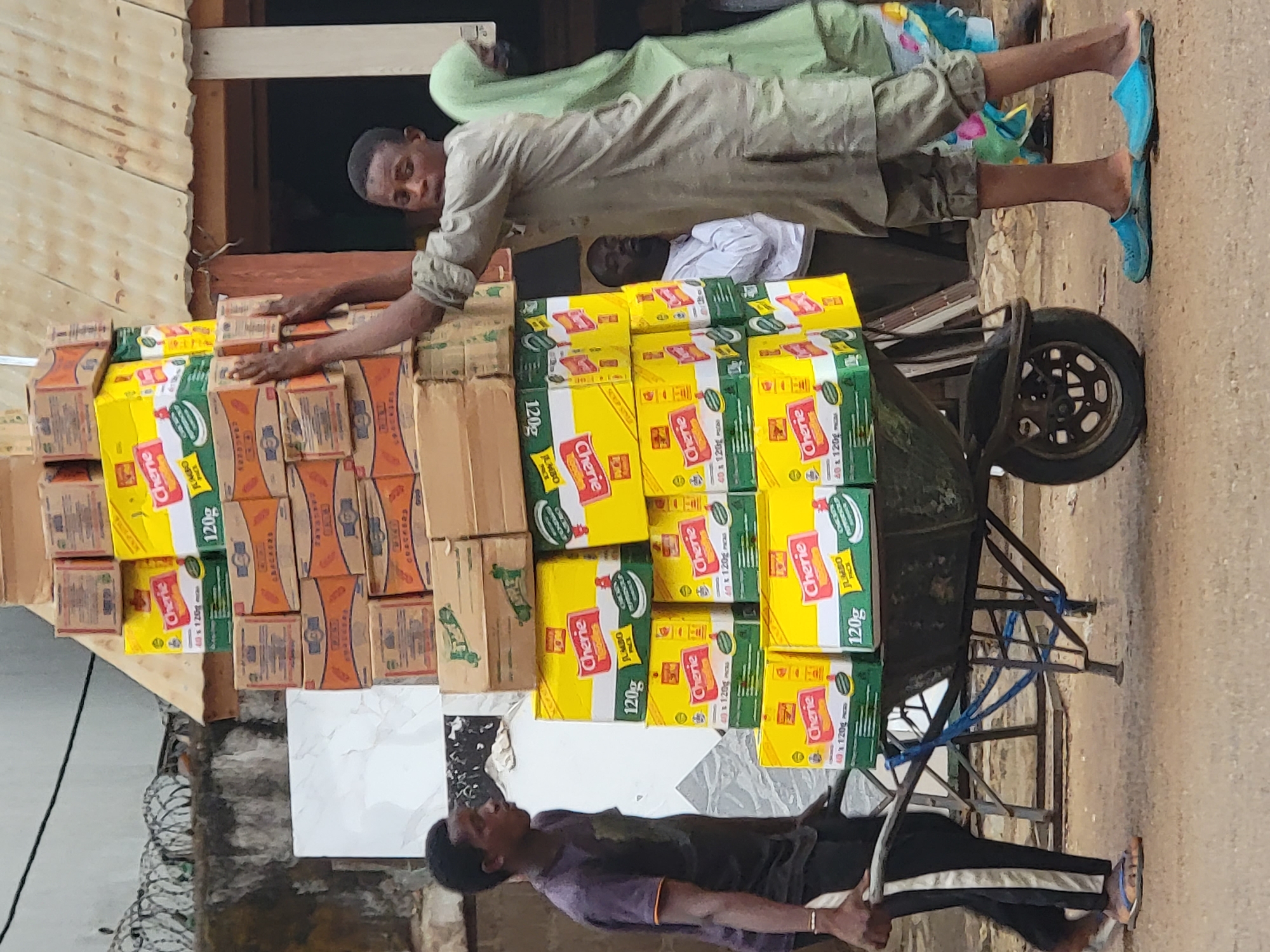 The image, taken in Maiburiji, Nigeria, captures an outdoor scene centered around the transportation of goods. A wheelbarrow is heavily laden with numerous cardboard boxes, forming a precarious stack. Many of these boxes are distinctly yellow and green, featuring the brand name "Cherie" prominently. Visible text on these boxes includes "120g," "JUMBO PACK," and "40 x 120g." Some boxes also display "SOUP MIX." Other boxes are plain brown.
In the foreground, a man wearing a dark t-shirt and track pants holds the handles of the overloaded wheelbarrow, appearing to be either preparing to move it or resting. Another man, dressed in a light-colored garment, is positioned beside a wooden structure, reaching over and securing or arranging the stacked boxes. A third individual is partially visible in the background. The ground appears unpaved, and a metal fence or barrier is visible in the lower left. The scene is brightly lit, suggesting it is daytime. The overall impression is of a busy commercial activity taking place in a local market or street setting.