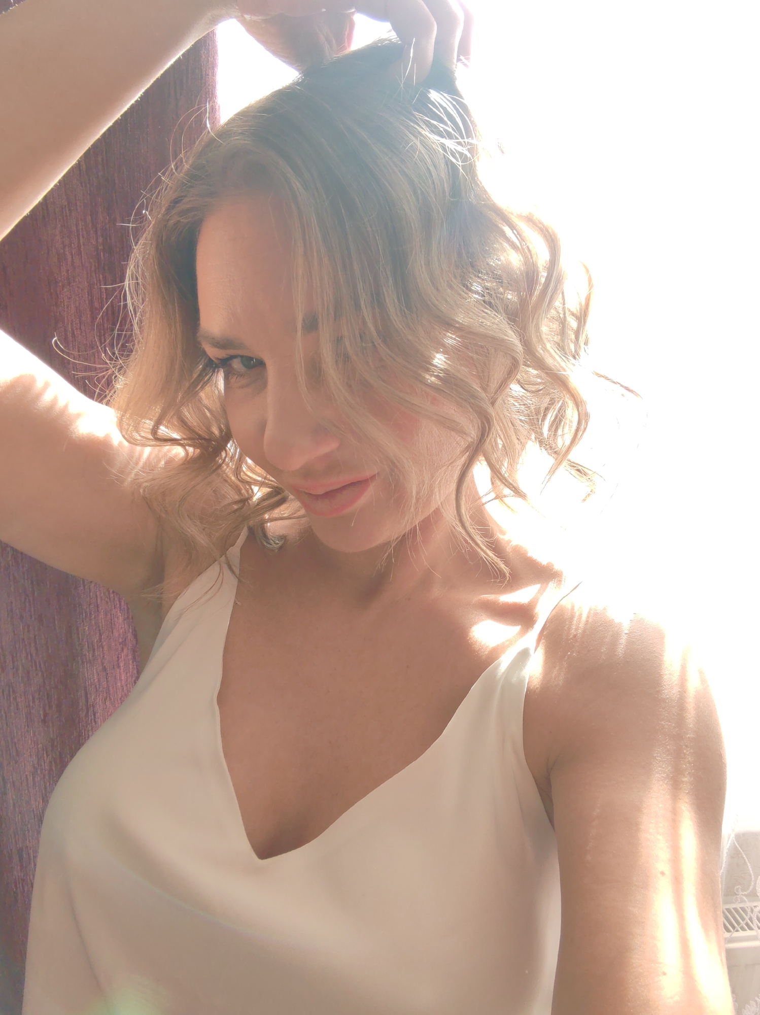 A brightly lit portrait features a woman with medium-length, wavy blonde hair, styled with loose curls. She is seen looking down and to her right with a gentle, slightly playful smile, her lips subtly parted. Her right arm is raised, with her hand seemingly touching or running through her hair. She is wearing a simple, light-colored (possibly white or off-white) V-neck camisole or top.
The image is characterized by strong natural backlighting, likely from a window to her right, which creates bright highlights on her hair and shoulders and some lens flare. The intense light obscures much of the background on the right side, but illuminates parts of her arm and décolletage. To her left, a dark, textured curtain, appearing purple or maroon, provides a contrasting backdrop. A small segment of a white decorative screen or fixture is visible in the bottom right corner. The powerful sunlight suggests a bright day in Saint Petersburg, Russia. No visible text is present. The overall mood is soft and warm, emphasizing the natural light and her expression.