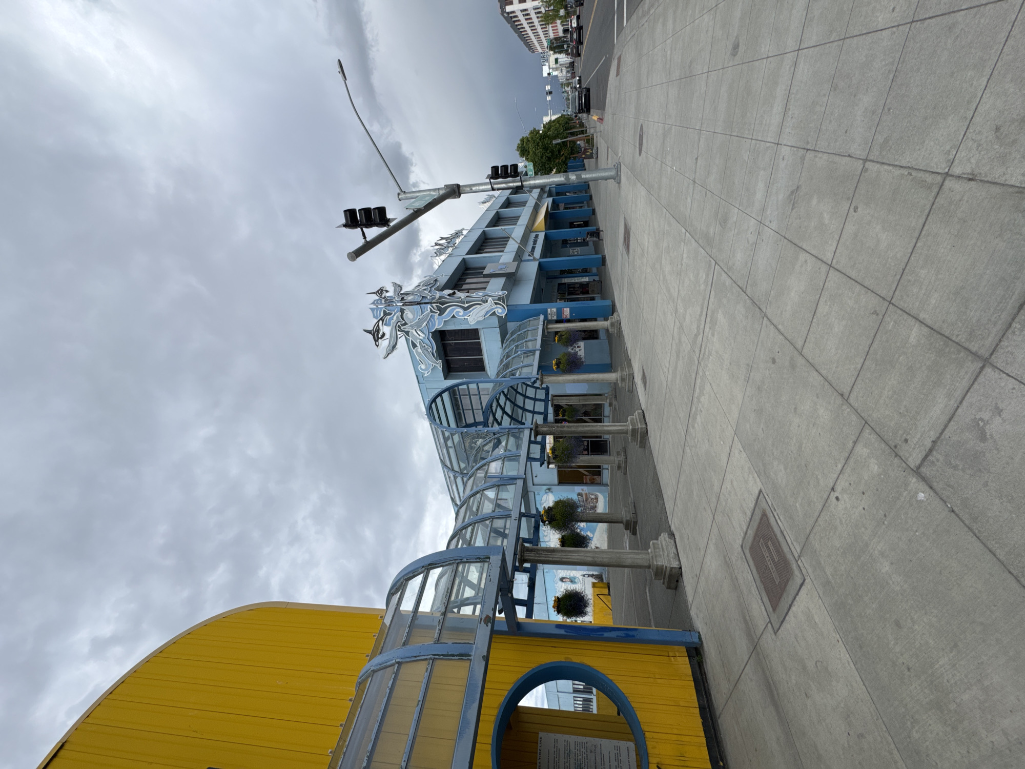 An outdoor, eye-level, wide-angle shot captures a cloudy day in downtown Anchorage, Alaska, USA. The foreground is dominated by a grey concrete sidewalk. To the left, a curved, bright yellow building with a blue trim features a circular cutout and an adjacent covered walkway with a glass roof and blue supports. The walkway leads to a blue building with a stylized white orca decoration on its facade. This building has multiple windows and doors. A sidewalk stretches ahead, leading to a street with a traffic light. Cars are visible on the street, moving away from the camera. The sky is overcast with grey clouds.