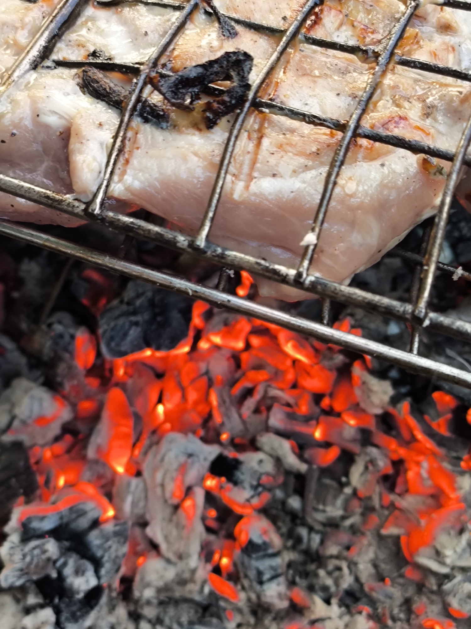 A close-up, top-down view shows pieces of meat on a metal grill over glowing red coals. The meat appears to be raw or partially cooked, with some charred bits visible. The coals are intensely hot and brightly lit, indicating they are actively burning. The scene is set outdoors, likely during a barbecue or grilling session. There are no people or specific city locations identifiable in the image.