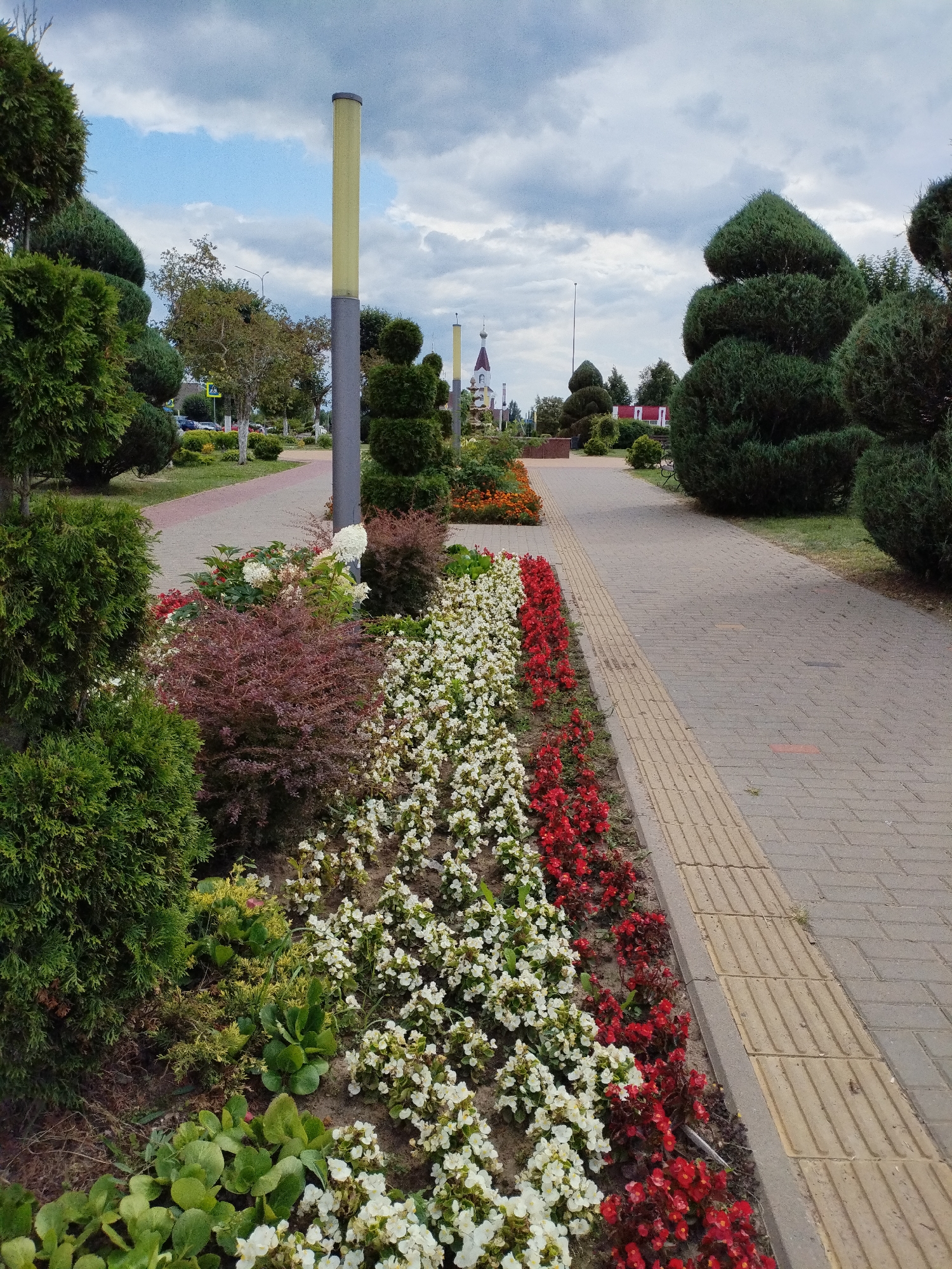 This is an outdoor, eye-level, wide shot of a paved walkway lined with flower beds and manicured bushes. The walkway is made of light gray rectangular pavers with a textured yellow strip on the right side, likely for the visually impaired. To the left of the walkway, a variety of plants are visible, including rows of white and red flowers, burgundy bushes, and large, cone-shaped green trees. In the background, a light pole with a yellow top stands near the walkway. Further in the distance, a church with a red roof and white spire can be seen, along with other buildings and trees. The sky is overcast with clouds. The scene is devoid of people. This appears to be a park or public garden area within the city of Bialynichy, Belarus, given the context.