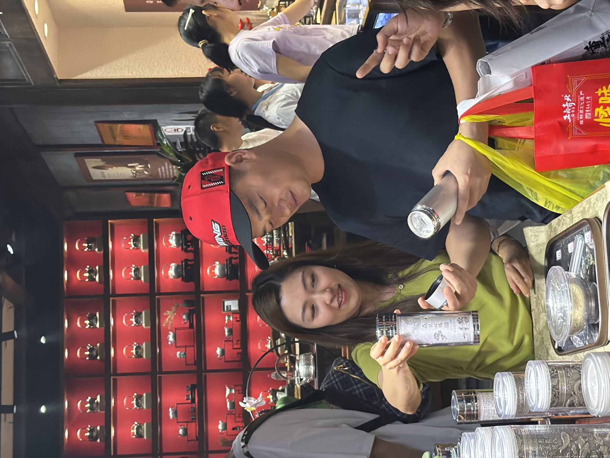 In the city of Yuzhong District, China, a man wearing a red baseball cap and a black t-shirt stands with a woman in a lime green shirt, both holding containers. The woman is holding an open cylindrical container with a white lid in her right hand and examining its contents. She is also holding another similar container in her left hand. The man is holding a silver cylindrical object in his right hand and has his left arm extended, with several shopping bags hanging from his wrist.
The background suggests a retail environment, likely a shop selling tea or related products. Red-backed shelves display numerous decorative items, possibly teapots, arranged in rows. The lighting is artificial and appears to be from overhead fixtures. Other shoppers are visible in the background, some blurry and out of focus, including a woman in a light purple t-shirt and a man in a white t-shirt. A plaid backpack is visible on the woman's left side. The overall scene depicts a moment of shopping and interaction within a commercial setting.