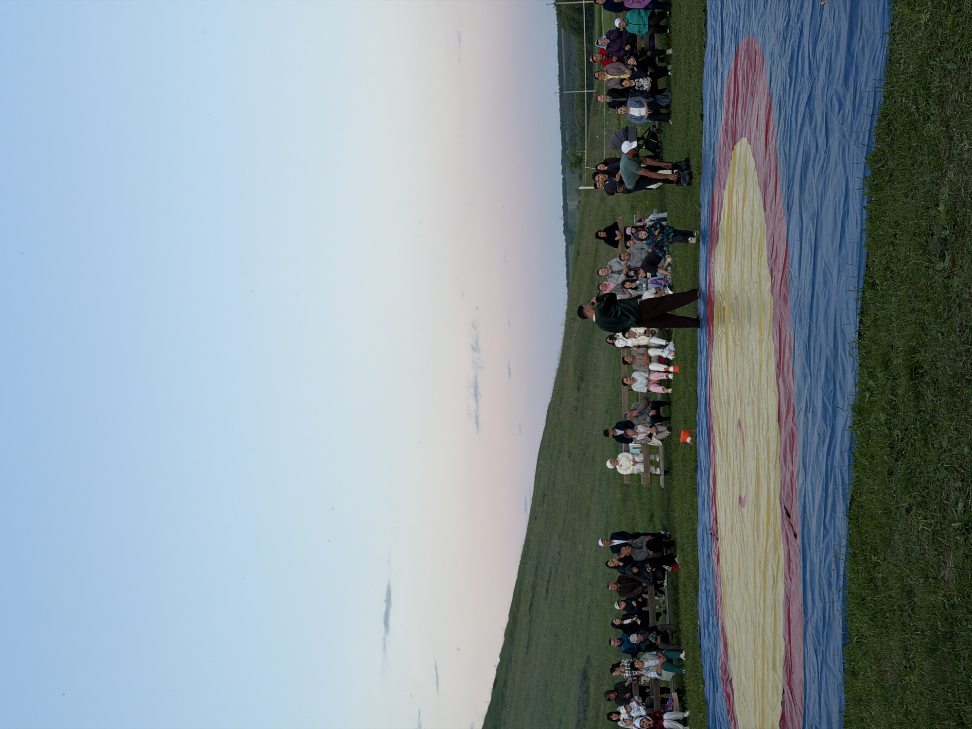 This image captures an outdoor gathering at an unknown location in Unknown, Unknown, likely during dusk or dawn due to the pastel blue and pink sky. In the foreground, a large, rectangular wrestling mat, primarily blue with a central yellow circle containing a red inner ring, lies on green grass. The mat is wrinkled. To the left of the mat, a man in a dark green shirt and dark trousers stands facing a seated audience, holding what appears to be a microphone. To the right of the mat, another man in a light-colored cap and light green shirt is bending over near the mat. A small child in a patterned jacket walks towards the bending man on the mat.
Approximately 30-40 individuals, varying in age from children to elderly, are seated on multiple wooden benches arranged in rows behind the mat, observing the activity. They are dressed in casual attire. An orange cone or small container is on the grass between the mat and the rightmost benches. Behind the spectators on the right, a baby stroller and a white net, possibly for volleyball, are visible. The background features a large, grassy green hill sloping upwards on the left, meeting the soft-hued sky. Distant trees and utility poles are visible on the horizon to the right.