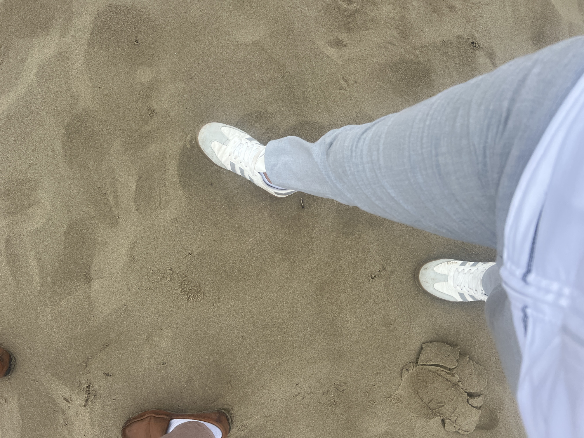 This high-angle, first-person perspective image captures a view of feet on sandy ground, likely a beach in Martil, Morocco. The main subject, positioned on the right, wears light grey pants and white sneakers with grey stripes. A white garment, possibly a shirt, is partially visible near the top right. To the bottom left, another person's feet are partially visible, wearing brown, low-cut shoes or slippers with white socks. The sand features ripples and textural variations, suggesting it may be slightly damp. A distinct, abstract sand formation or imprint, resembling a partial footprint or a small sand sculpture, is visible on the bottom right, next to the main subject's left foot. The lighting is soft and even, indicative of daytime without harsh shadows. No text is discernible in the image. The scene conveys a moment of standing or walking on the sandy coast.