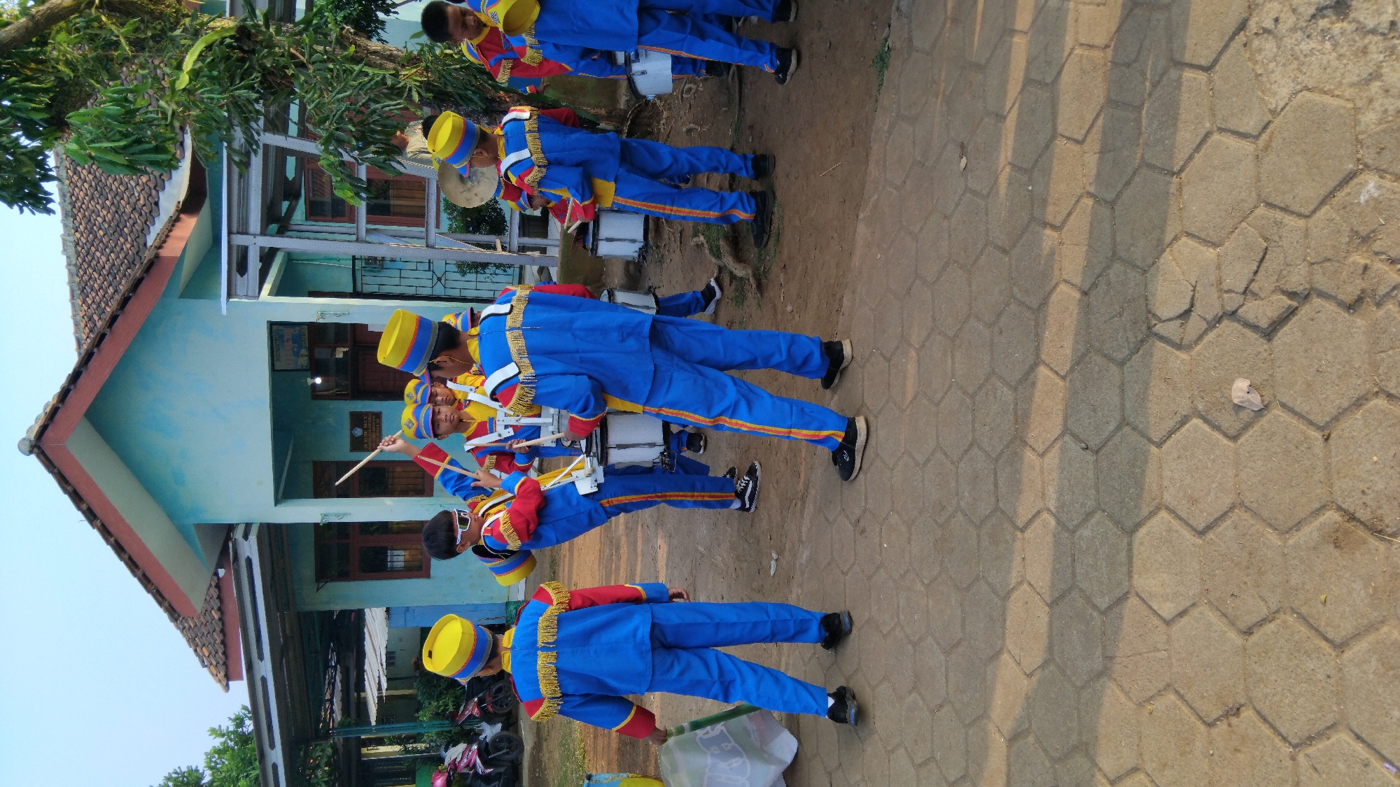 In Sembojo, Indonesia, a group of children in blue and yellow marching band uniforms are gathered outside a light blue building with a brown tiled roof. The children are holding drums and drumsticks, with some actively playing. One child in the foreground, facing away from the camera, wears a blue uniform with yellow trim and a yellow hat, holding a green flag pole. Another child, also in a similar uniform, is positioned to the left and slightly behind, facing the building. The ground is paved with hexagonal interlocking concrete blocks. A motorcycle is parked to the left of the building. The scene is outdoors, under a clear sky, with vegetation visible around the building.