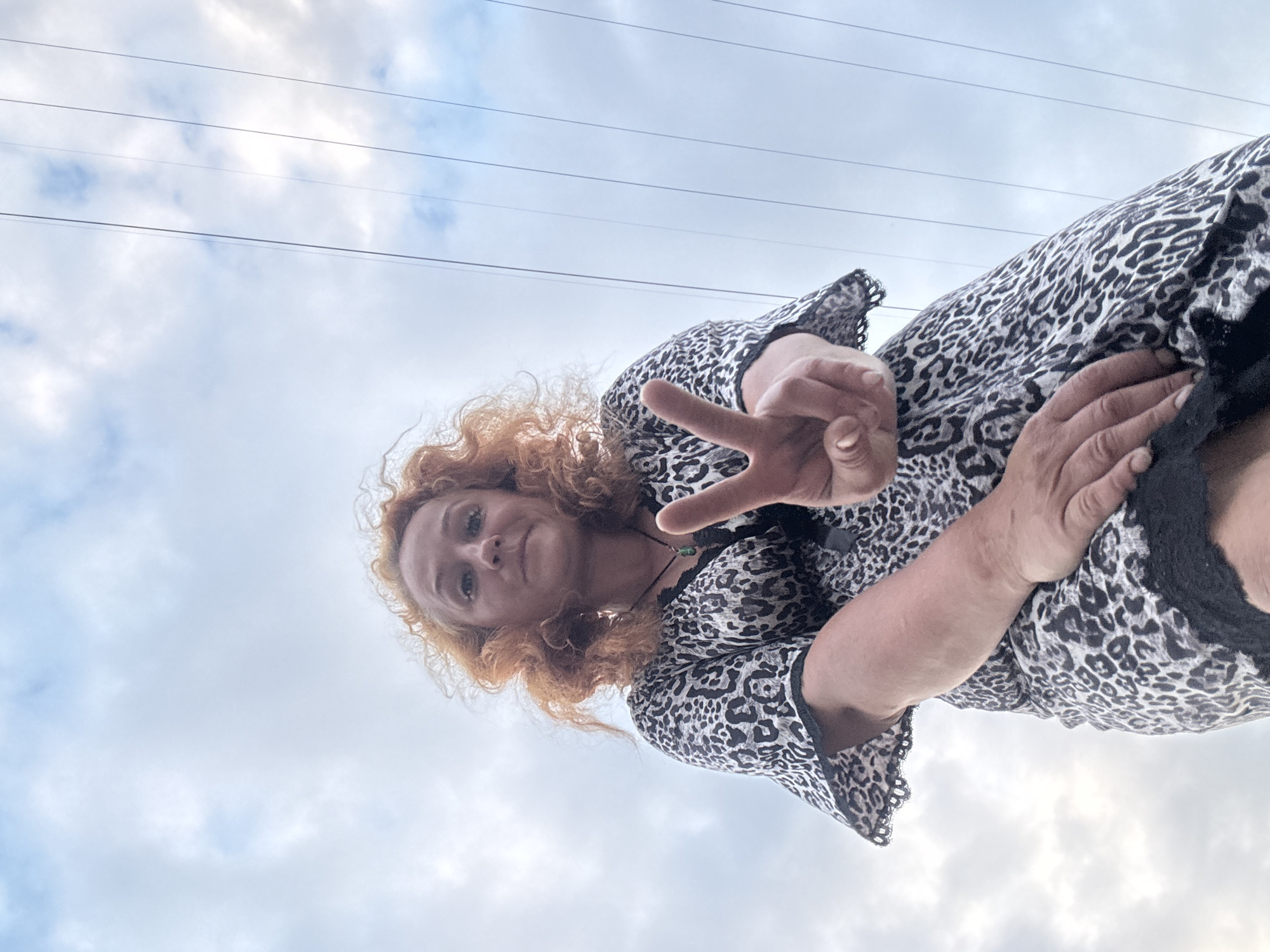 A low-angle, wide shot shows a woman with curly, auburn hair looking up at the camera. She is wearing a black and white leopard-print dress with black lace trim. Her right hand is raised, fingers forming a peace sign. Her left hand is visible near her chest. The background is a cloudy sky with power lines stretching across it. The image appears to be taken outdoors during the day, with the lighting suggesting it could be late afternoon or early evening. The overall mood is casual and relaxed. There is no visible text in the image.