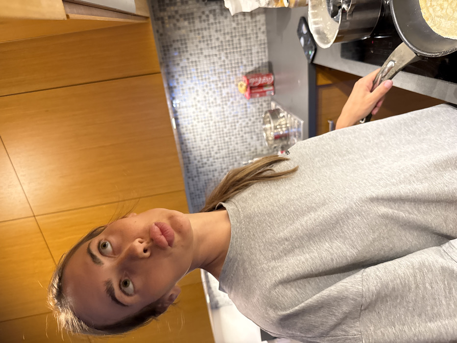 A female individual is depicted in a kitchen setting in Warsaw, Poland, engaged in cooking. She possesses light skin, light-colored eyes, and long brown hair secured in a ponytail. Her head is tilted slightly upwards, and her gaze is directed towards the upper left, while her lips are pursed in a pout. She wears a grey, short-sleeved, crew-neck t-shirt. Her right hand, partially visible, grips the handle of a dark frying pan situated on a black electric stovetop. Inside the pan, a light-colored, circular food item, possibly a pancake or crepe, is visible.
The kitchen environment features light brown wooden upper cabinets and a grey mosaic tile backsplash with under-cabinet lighting. A dark grey countertop is present, upon which several items are arranged: two red Coca-Cola cans, a silver pot or bowl, and a black smartphone lying face down. Another metallic pot is partially visible near the frying pan on the stovetop. The scene is illuminated by artificial indoor lighting.