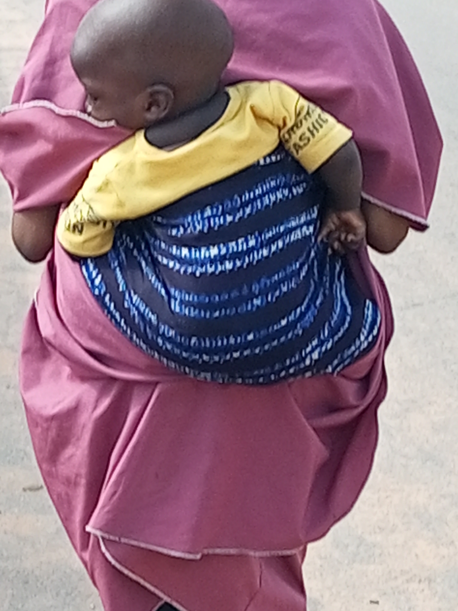 The image shows a close-up, rear view of a person carrying a baby on their back. The person is wearing a pink garment, possibly a dress or wrapper. The baby is wearing a yellow shirt with dark blue and white striped fabric wrapped around its waist and the carrier's torso. The baby's head is visible, and it appears to be looking downwards. The scene is outdoors, with a blurred, light-colored ground, suggesting a street or pathway. The image is cropped tightly, focusing on the individuals and their attire. The general context of Gombe, Nigeria, implies an urban or semi-urban environment where such carrying practices are common.