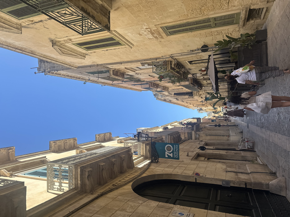 The image captures a narrow, stone-paved street in the historic center of Lecce, Italy, identified as Via Libertini. The architecture consists of multi-story buildings constructed from light-colored stone, featuring Baroque elements such as ornate wrought-iron balconies and windows with louvered shutters. On the left, a large dark arched wooden door marks the ground floor of a building from which a teal banner displaying "70 UNI SALENTO" hangs. The scene is under clear daylight with a bright blue sky, casting defined shadows.
Multiple individuals are visible walking away from the viewer down the street. In the foreground, two females are prominent: one wearing a light-colored dress and carrying a white shoulder bag, the other in patterned trousers and a white t-shirt with a multi-colored tote bag. Other figures are present further down the street, one of whom wears a long, dark-colored garment. On the right side of the street, an outdoor dining area is set up with dark tables, chairs, and large umbrellas, flanked by tall potted green plants. A white plastic chair rests against the wall on the left side of the street, near a partially visible traffic sign.