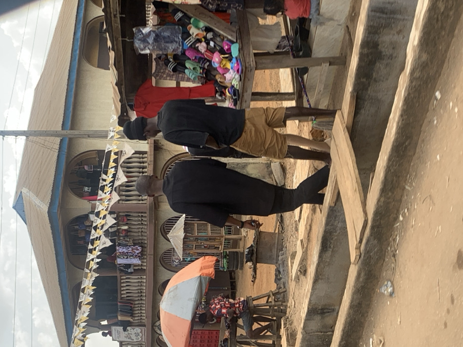 This image depicts an outdoor market scene in Egbedi, Nigeria, characterized by unpaved ground and commercial activity under a cloudy sky. In the foreground, a wide concrete gutter or ditch traverses the scene, with two wooden planks serving as makeshift bridges. Two adult males are walking away from the viewer, traversing the area near the ditch. The man on the left wears a dark tunic over dark trousers. The man on the right wears a black t-shirt, tan shorts, and a black beanie, with a light-colored object visible on his left foot.
To the far left, a man in a patterned shirt and shorts is seated on a wooden stool at a wooden table, partially shaded by an orange and white umbrella. Near him, a dark metal basin rests on the ground. To the far right, a small child in a red top sits on a low concrete ledge. Adjacent to the child, a market stall displays numerous colorful sandals and slippers on a wooden tiered stand, with a red garment hanging behind it.
In the background, a two-story building with a brown corrugated metal roof and blue trim dominates the upper half of the image. The upper floor features a balcony with a white balustrade, from which various clothes are hung. Yellow and black patterned bunting is strung across the balcony. Several arched openings on both floors reveal more merchandise, suggesting shops within the building. Other individuals are visible on the balcony and further within the market.