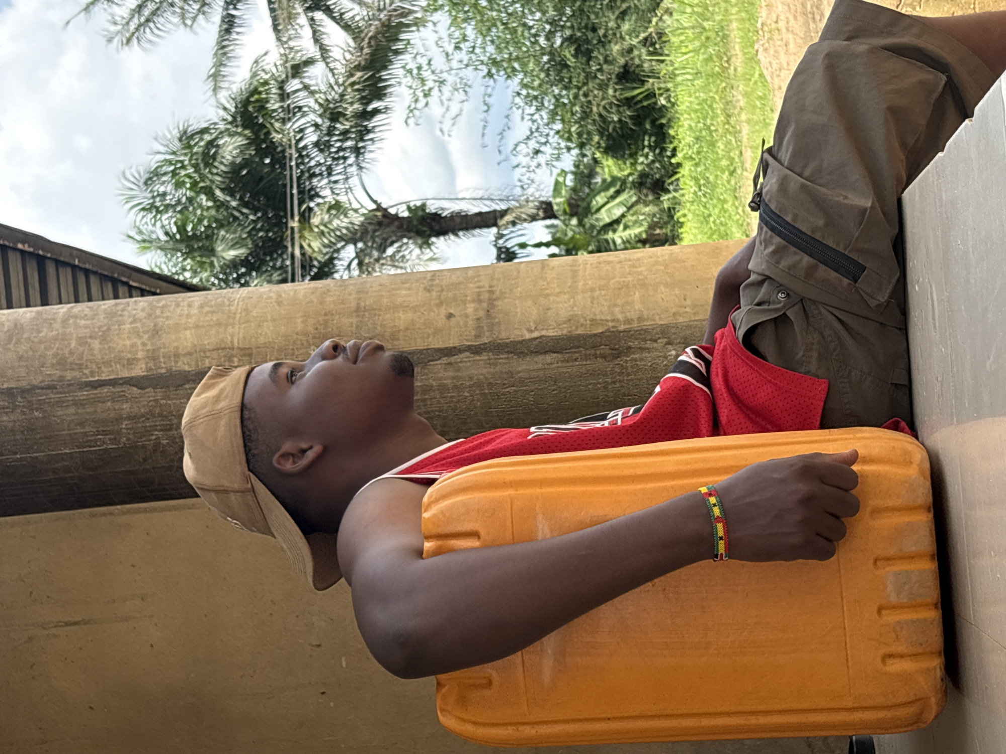 A man is lying on his back in an outdoor environment in Ikot Ekpene, Nigeria. He is wearing a tan baseball cap, a red and white basketball jersey, and khaki shorts. He is using a yellow rectangular container as a pillow, with his arms crossed over it. A beaded bracelet is visible on his left wrist. Palm trees and other foliage are in the background, with a cloudy sky visible above. The man is positioned next to a weathered concrete structure.