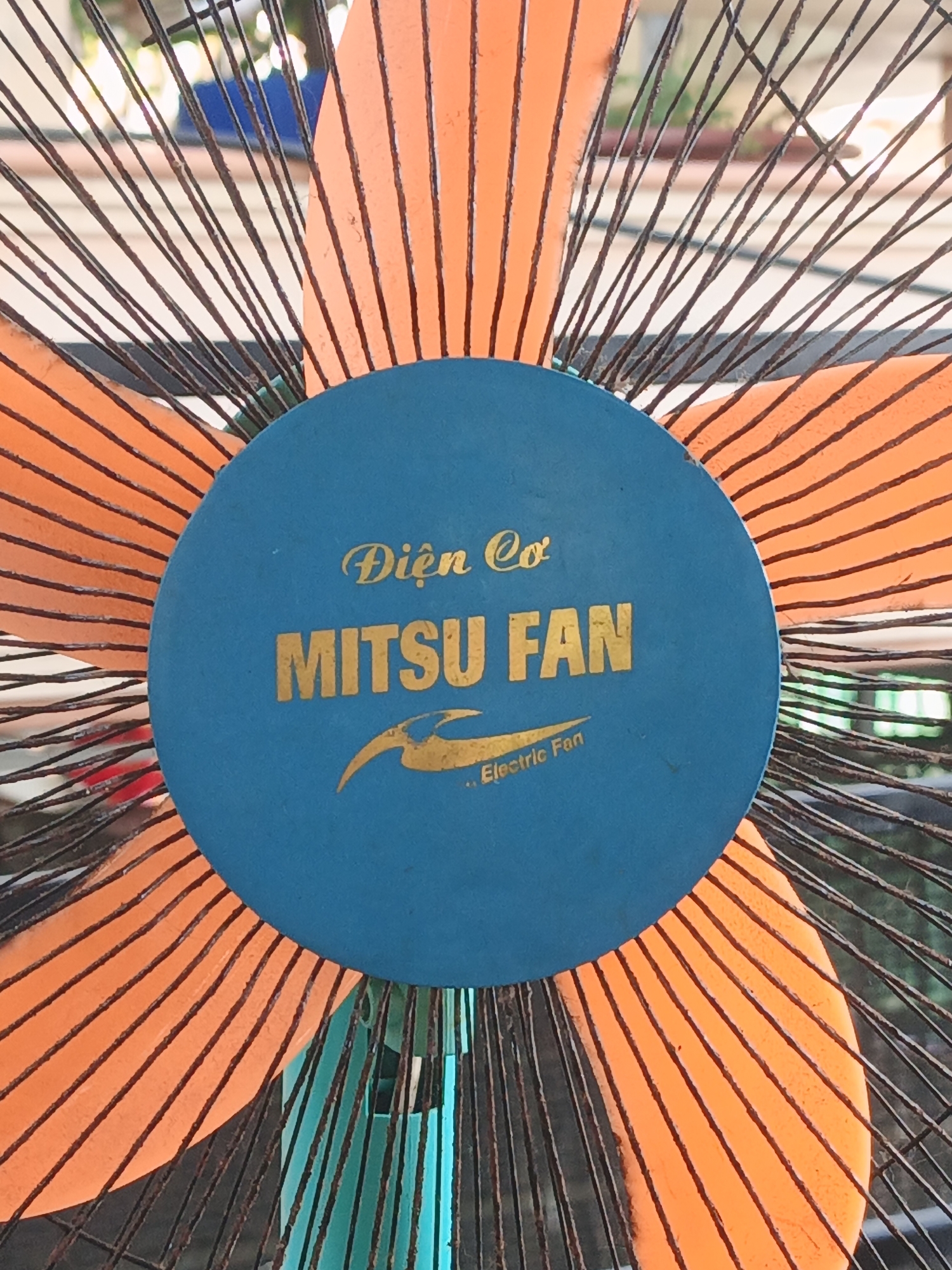 A close-up, eye-level shot shows the center of an electric fan with orange blades and a blue center cap. The cap has gold lettering and a gold logo that appears to be a stylized bird or flame. The fan is outdoors, and the background consists of blurred foliage and what appears to be a building. The fan is stationary. The location is the city of Bac Nha Trang Ward, Vietnam.
