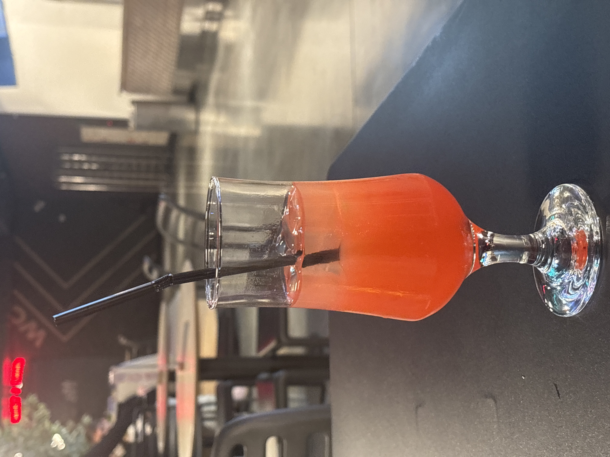 A tall glass with a vibrant orange beverage and a black straw stands on a dark, textured surface. The glass has a slender stem and a wide, flared opening. The drink appears to be a cocktail with ice. In the background, out of focus, are elements of what appears to be an indoor public space. To the left, red neon lights are visible above a sign that reads "WC". Further back, tables and chairs are arranged, suggesting a cafe or restaurant setting. To the right, there are vertical metallic structures and a cushioned bench, possibly part of the seating area. The overall lighting is dim, with reflections on the surfaces indicating ambient light sources. The image appears to be taken in Saint Petersburg, Russia, based on the presence of the "WC" sign, which is commonly used in Russian-speaking countries.