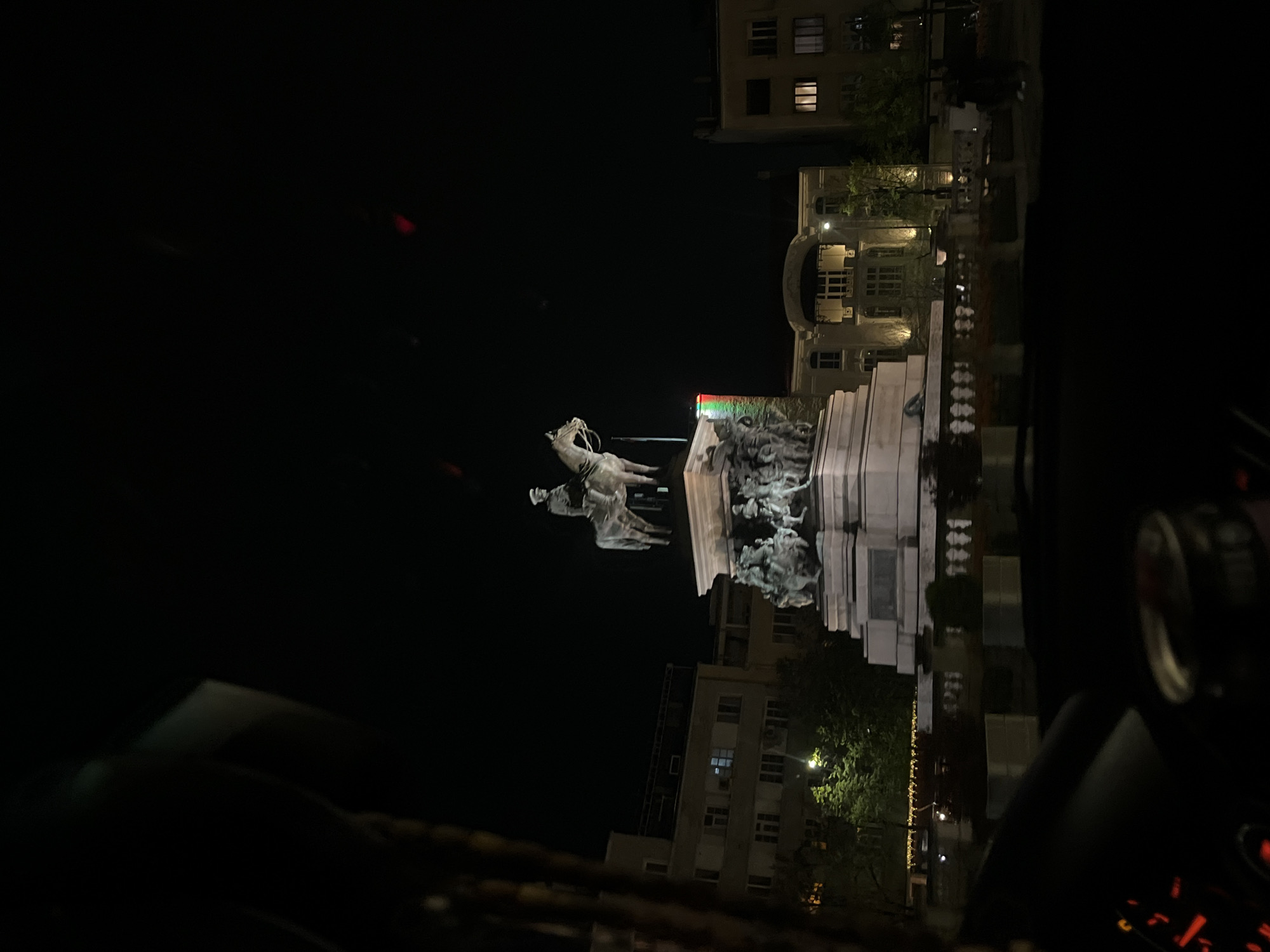 A nighttime, medium-shot image shows the Equestrian statue of Tsar Alexander II on the Liberation Monument in Sofia, Bulgaria. The monument is illuminated and features a multi-tiered base with sculptures. Buildings are visible in the background, lit by streetlights. The photograph appears to have been taken from inside a vehicle.