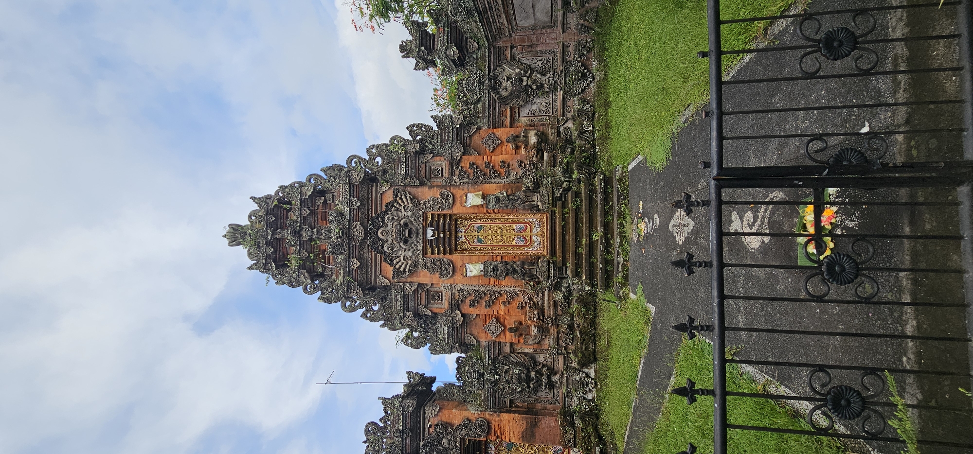 A ground-level, medium shot depicts the exterior of a Balinese temple structure located in the Ubud District, Indonesia. The temple is made of red brick and is decorated with intricate stone carvings. It is topped with a series of tiered roofs. The temple is surrounded by lush green vegetation. A black wrought-iron gate is situated in the foreground, partially obscuring the temple. A small offering of flowers on a green leaf is placed behind the bars of the gate. The sky is visible in the upper left corner of the image, it is blue with white clouds. The perspective is slightly tilted upward.