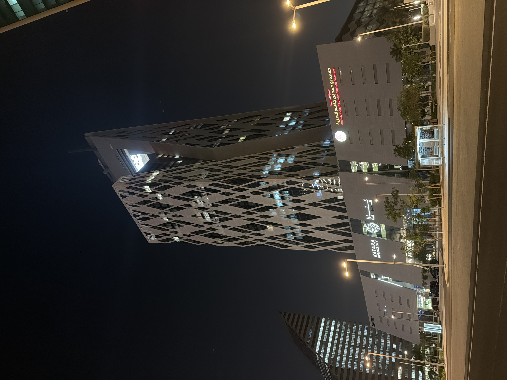 The image captures a modern urban night scene in Lusail, Qatar, showcasing distinctive contemporary architecture under a dark, clear sky. The central focus is a tall, geometrically designed skyscraper with a striking facade featuring a crisscross or diamond pattern, reflecting lights from its interior and surroundings. This prominent building also appears to have a large, illuminated cut-out section near its top.
To the right of this skyscraper stands a rectangular, multi-story building, which houses the "Jassim & Hamad Bin Jassim Charitable Foundation," clearly marked in both Arabic ("مؤسسة جاسم و حمد بن جاسم الخيرية") and English, along with a circular logo. Further down and to the right, another building is visible, bearing the sign "KATARA HOSPITALITY" and an associated circular logo, possibly depicting a tree. On the far bottom left, another skyscraper with a more conventional grid-like window pattern is partially visible, with the word "LOTUS" displayed vertically on its facade.
The scene is illuminated by numerous streetlights, casting a warm glow on the surroundings. A wide road or paved area runs along the right side of the image, bordered by scattered trees and small bushes. A small, illuminated structure, possibly an information kiosk or bus stop, is situated on the pavement. No people are visible, lending a quiet, serene atmosphere to the bustling urban landscape. The overall impression is one of modern development and sophisticated urban planning.