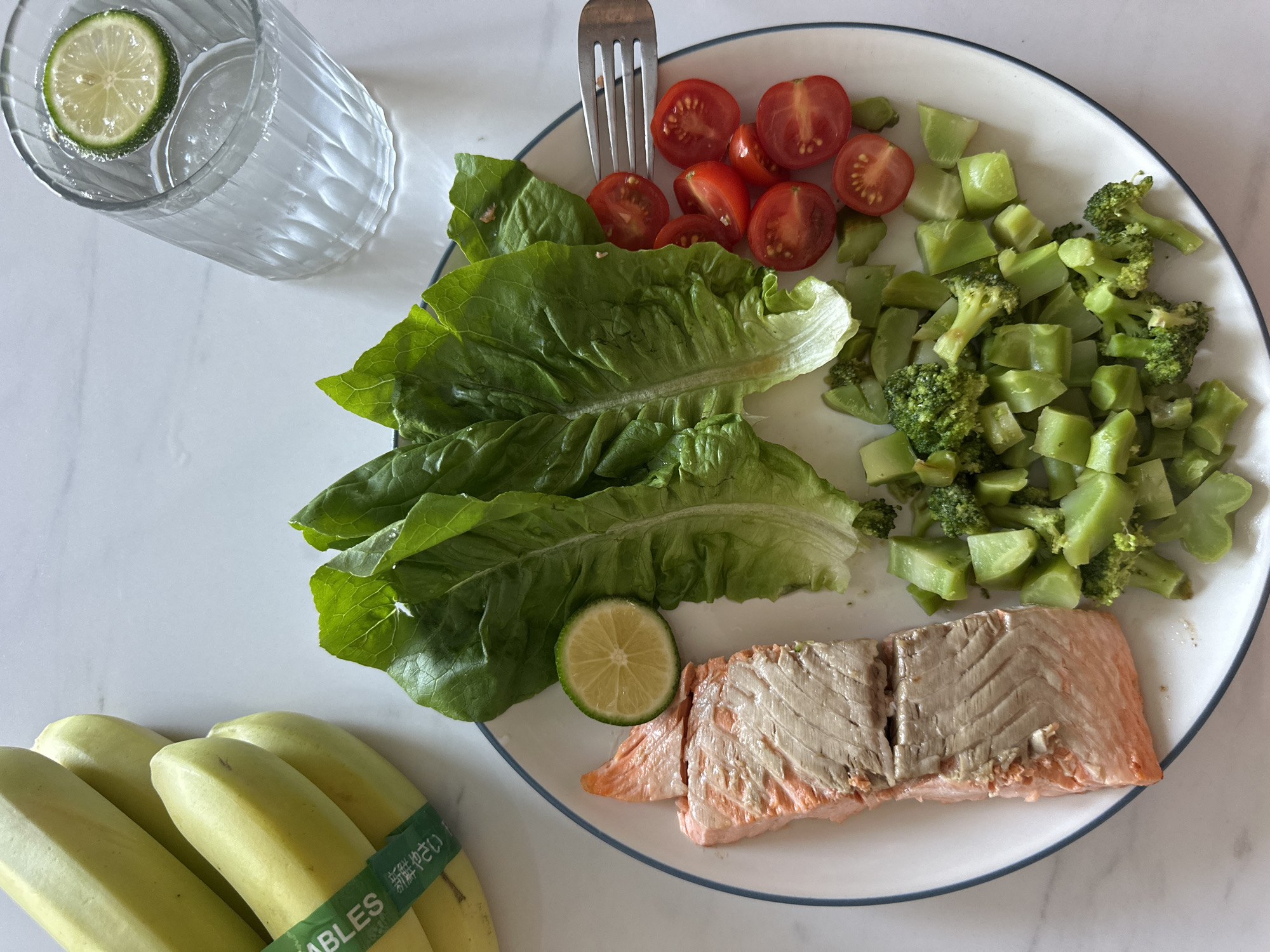 The image shows a plate of food and a glass of water with lime, on a white marble surface. The plate contains lettuce, chopped tomatoes, chopped broccoli, a lime slice, and salmon. There is a fork piercing the tomatoes. To the left, are a bunch of bananas. The background is out of focus. The image was taken in Mengwi, Indonesia.
