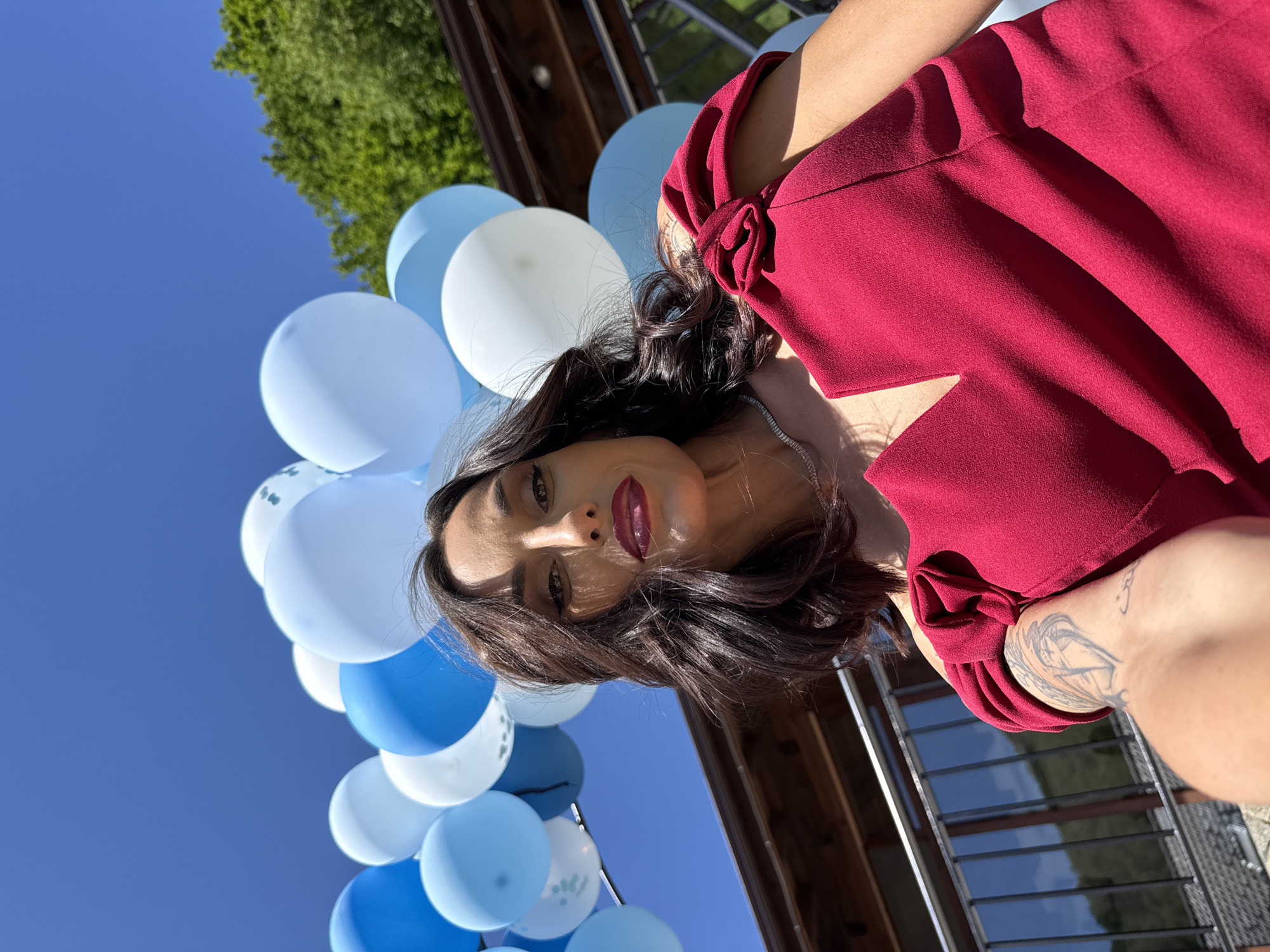 A woman in a burgundy dress is positioned to the right of a bunch of blue and white balloons. The scene is outdoors under a bright blue sky. The location is Ribaritsa, Bulgaria. Behind the balloons, there is a wooden structure and some green foliage. The woman is looking directly at the camera. The woman has dark hair.