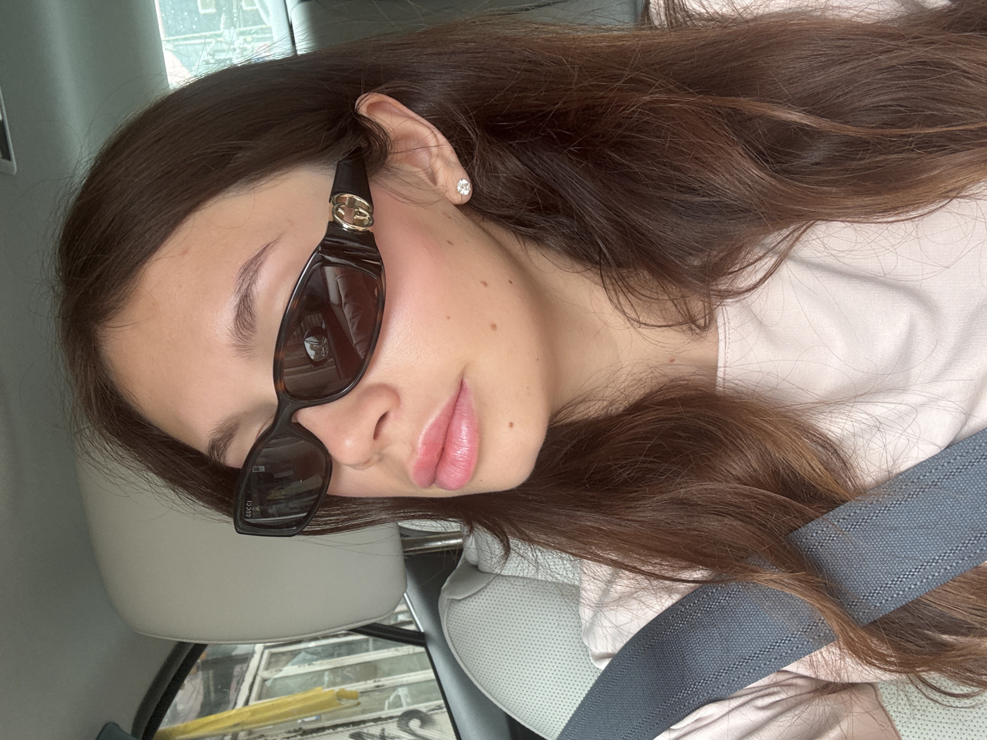 A young woman with long brown hair is in a car, wearing sunglasses and a seatbelt. She is facing the camera with her head tilted. The environment appears to be the interior of a vehicle, with a grey seat and window visible. There is no clear indication of the city of Tel-Aviv, Israel, in the image.