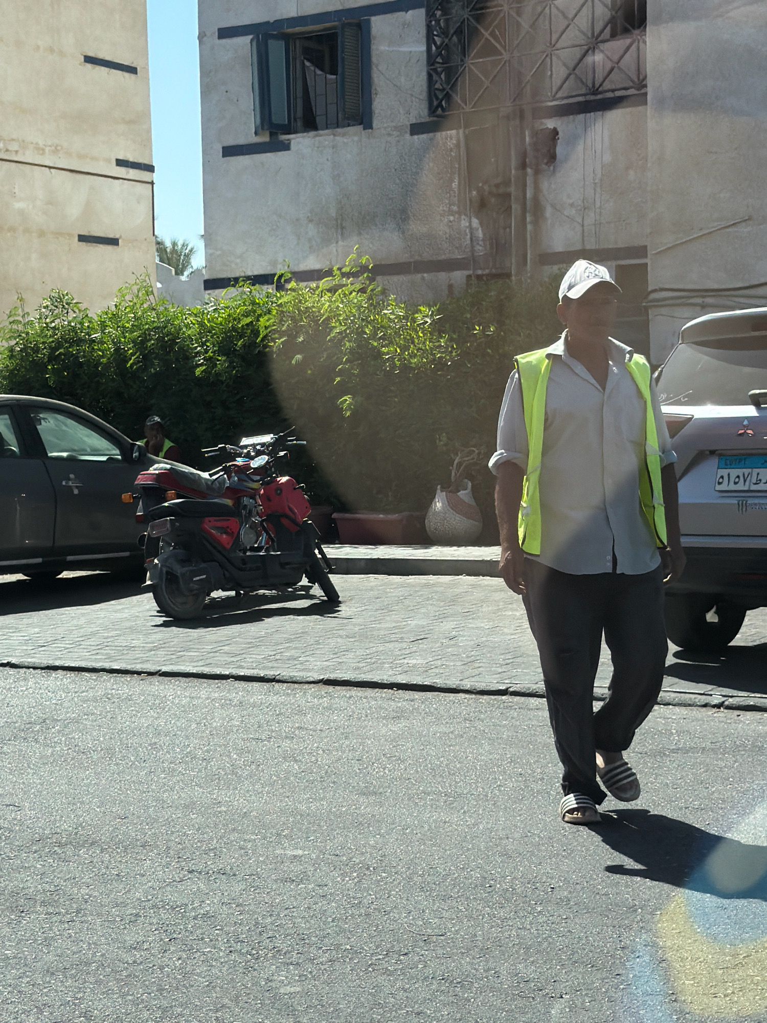 A man wearing a white cap, a light-colored collared shirt, a bright yellow vest, and dark pants walks on a paved street in Sharm El Sheikh, Egypt. He is wearing striped sandals. To his left is a red and black scooter. Behind the scooter is a dark-colored car. In the background, there are buildings with windows and some greenery. A silver car with an Egyptian license plate is parked on the right side of the image. The sun is casting shadows on the ground.
