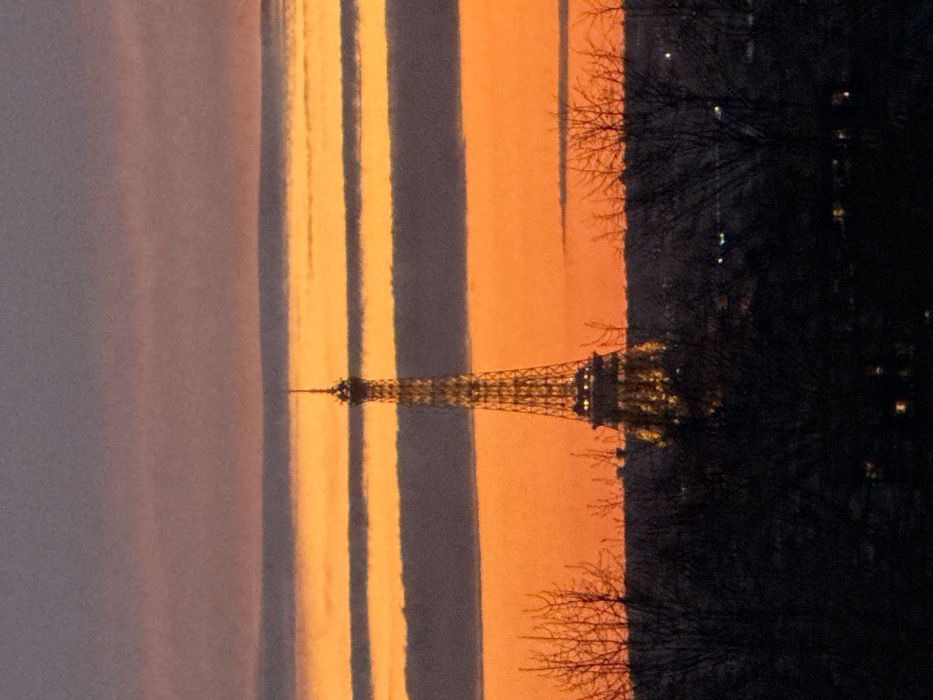 The image depicts the illuminated Eiffel Tower in Paris, France, against a vibrant sunset sky. The sky features distinct horizontal bands of color: deep orange dominates the lower and middle sections, interspersed with horizontal strips of dark grey and purple clouds, while the top portion of the sky is predominantly purple-grey. The Eiffel Tower, located in the 7th arrondissement of Paris, stands centrally in the mid-ground, brightly lit with warm yellow lights, highlighting its intricate lattice structure. Its spire extends into the upper sky.
In the foreground, a dark silhouette of bare tree branches frames the lower part of the scene, obscuring a portion of the distant cityscape. Below the trees, a dark horizon line shows scattered, faint points of light, indicative of city illumination from an elevated perspective. No discernible people, specific actions, or direct interactions are visible. The overall scene is a distant, elevated view of the iconic landmark at dusk or twilight.