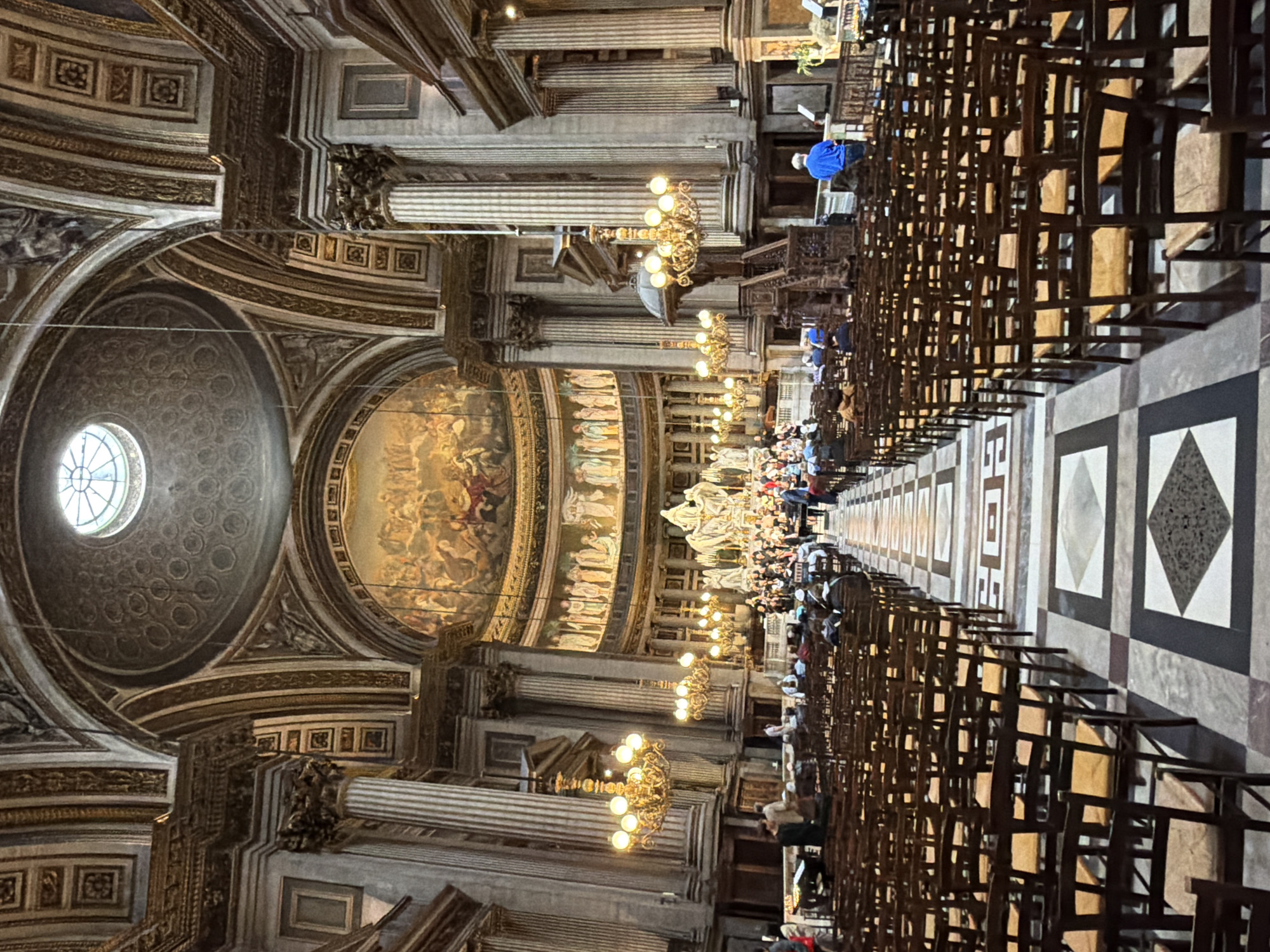 This is an image of the interior of the Madeleine Church in Paris, France. The shot is taken from the rear of the nave, looking towards the altar. The church features a grand dome with a skylight, ornate frescoes adorning the ceiling and apse, and tall Corinthian columns. Rows of empty wooden chairs with light-colored seats fill the nave, arranged on a patterned marble floor. In the distance, at the front of the church, a choir and orchestra are visible, performing in front of the altar, which is decorated with sculptures and religious iconography. Several people are seated in the chairs, and a few individuals stand near the choir and orchestra. The lighting is dim and warm, emanating from chandeliers and the skylight.