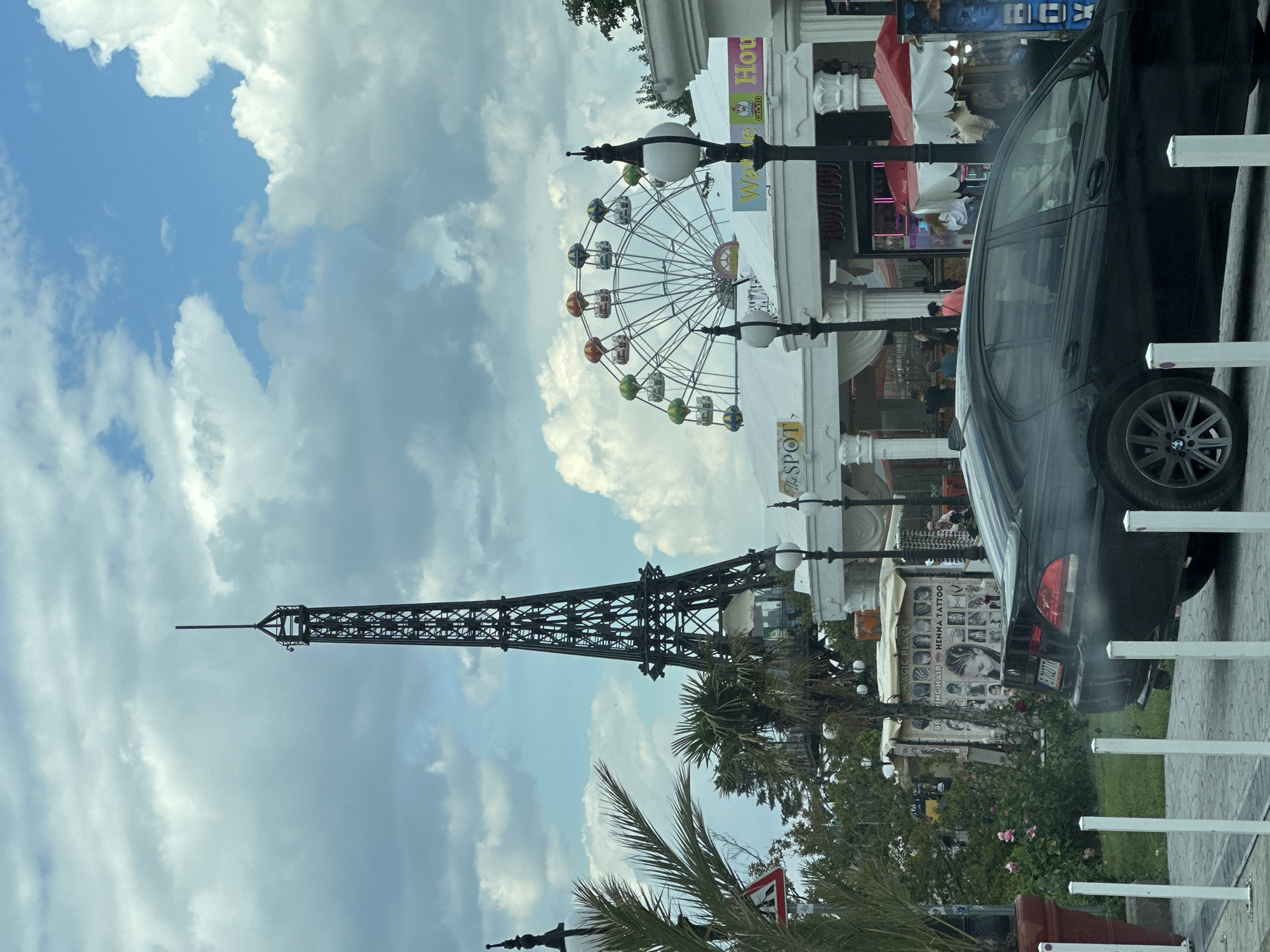 The image captures a scene in what appears to be a tourist area, likely Varna, Bulgaria, given the license plate of the vehicle. An Eiffel Tower replica stands tall against a backdrop of a partly cloudy sky. Nearby, a colorful Ferris wheel adds to the atmosphere. The scene is framed by palm trees, street lamps, and a sidewalk with white bollards. A black car partially obscures the view, and in the background, there are commercial establishments, one displaying henna tattoo designs. People are visible around the establishments.