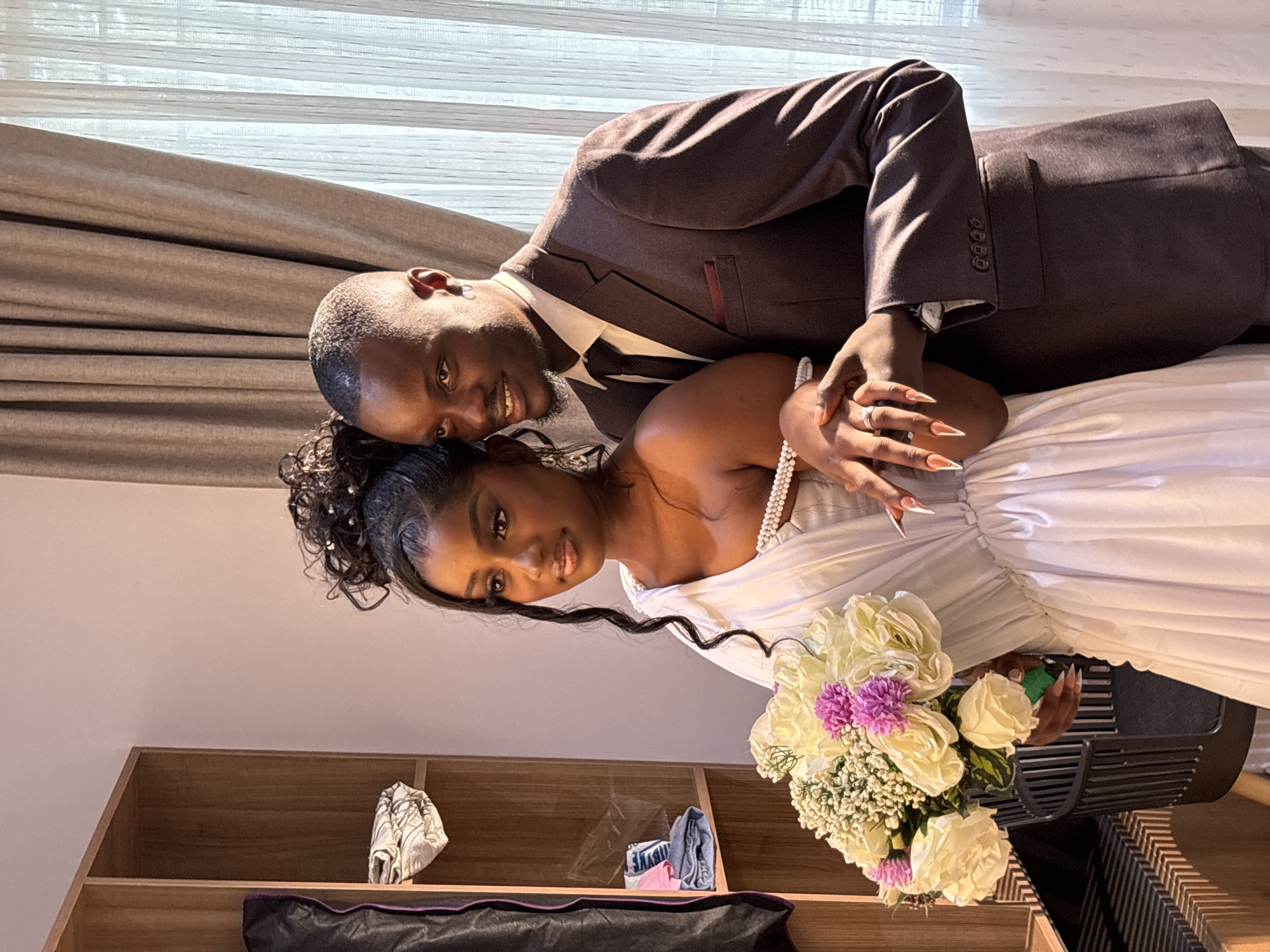 The image depicts a couple in Gura Top, Nigeria, likely on their wedding day. The man, dressed in a brown suit with a white shirt and tie, is leaning over the woman, who is wearing a white wedding dress and holding a bouquet of white and purple flowers. They are both smiling and looking towards the camera. The woman's hands are interlocked, and she is wearing a ring. In the background, there is a window with a curtain and a wooden cabinet with clothes and a black bag inside.