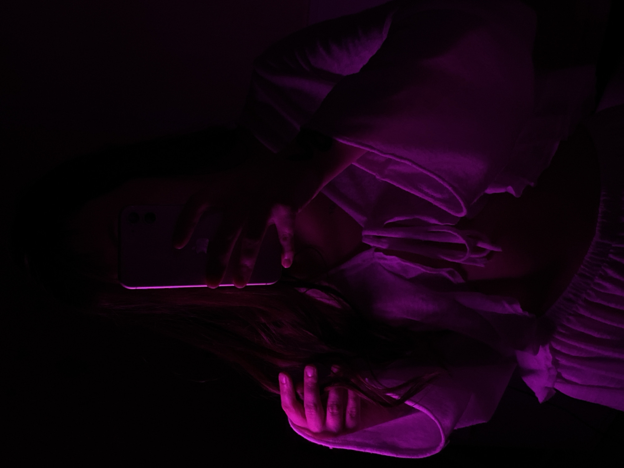 A person lies on their back, holding a smartphone above their face in a dimly lit environment with purple lighting. The person's face is obscured by the phone, and their long dark hair spreads around them. They are wearing a white blouse or shirt. No information about Votkinsk, Russia, can be derived from the image.