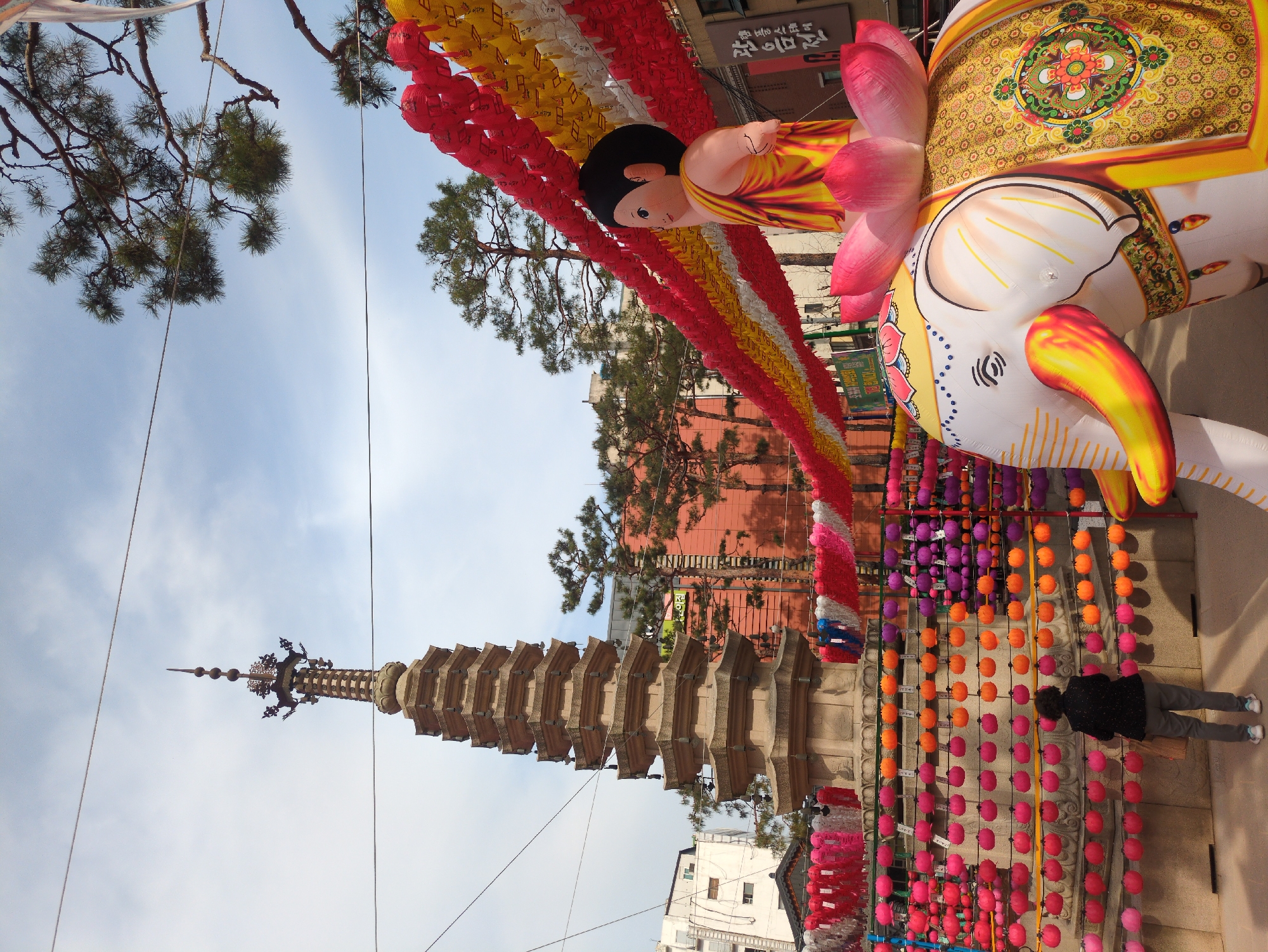 The image shows a pagoda and a festive scene, most likely captured in Seoul, South Korea, during a Buddhist celebration.
The prominent feature is a tall, multi-tiered stone pagoda. The pagoda is surrounded by strings of colorful lanterns, with red, orange, purple, and white predominating. The decorations suggest a Buddhist festival, most likely celebrating Buddha's Birthday.
To the right of the pagoda stands an inflated figure of a child Buddha seated on a lotus flower, riding atop an elephant.
A person is standing near the pagoda, looking at the lanterns.
The sky is blue with light clouds, suggesting a clear day. Buildings are visible in the background, typical of an urban area. Trees are scattered throughout the scene. Powerlines run through the image.