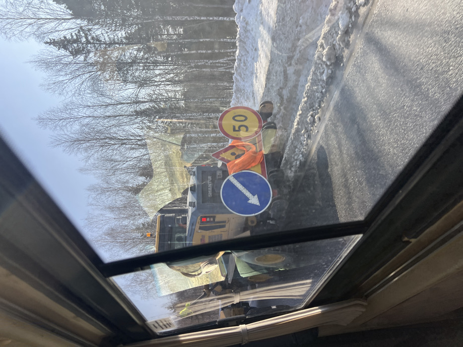 This image captures an outdoor scene in Pesterevo, Russia, viewed through the window of a vehicle. The perspective includes a window frame and part of a curtain on the left. The central subject is a yellow and gray 'SHANTUI' branded heavy construction vehicle, identifiable as a front-end loader or road clearing equipment, positioned on the right shoulder of an asphalt road. The vehicle displays an amber rotating beacon on its roof and illuminated red lights on its rear. Affixed to its rear are a blue circular mandatory sign with a white diagonal arrow pointing downward-left, a yellow triangular warning sign (partially obscured), and a yellow circular speed limit sign indicating "50". An orange high-visibility safety vest is draped over the equipment and signs.
The vehicle is actively engaged in pushing or clearing snow and slush from the road surface, creating distinct patterns of disturbed snow along the roadside. To the immediate right of the road, a substantial bank of accumulated snow is visible. The environment consists of a forest in the background, primarily composed of bare deciduous trees with some interspersed evergreen conifers, set against a clear blue sky. No individuals are explicitly visible within the frame.
